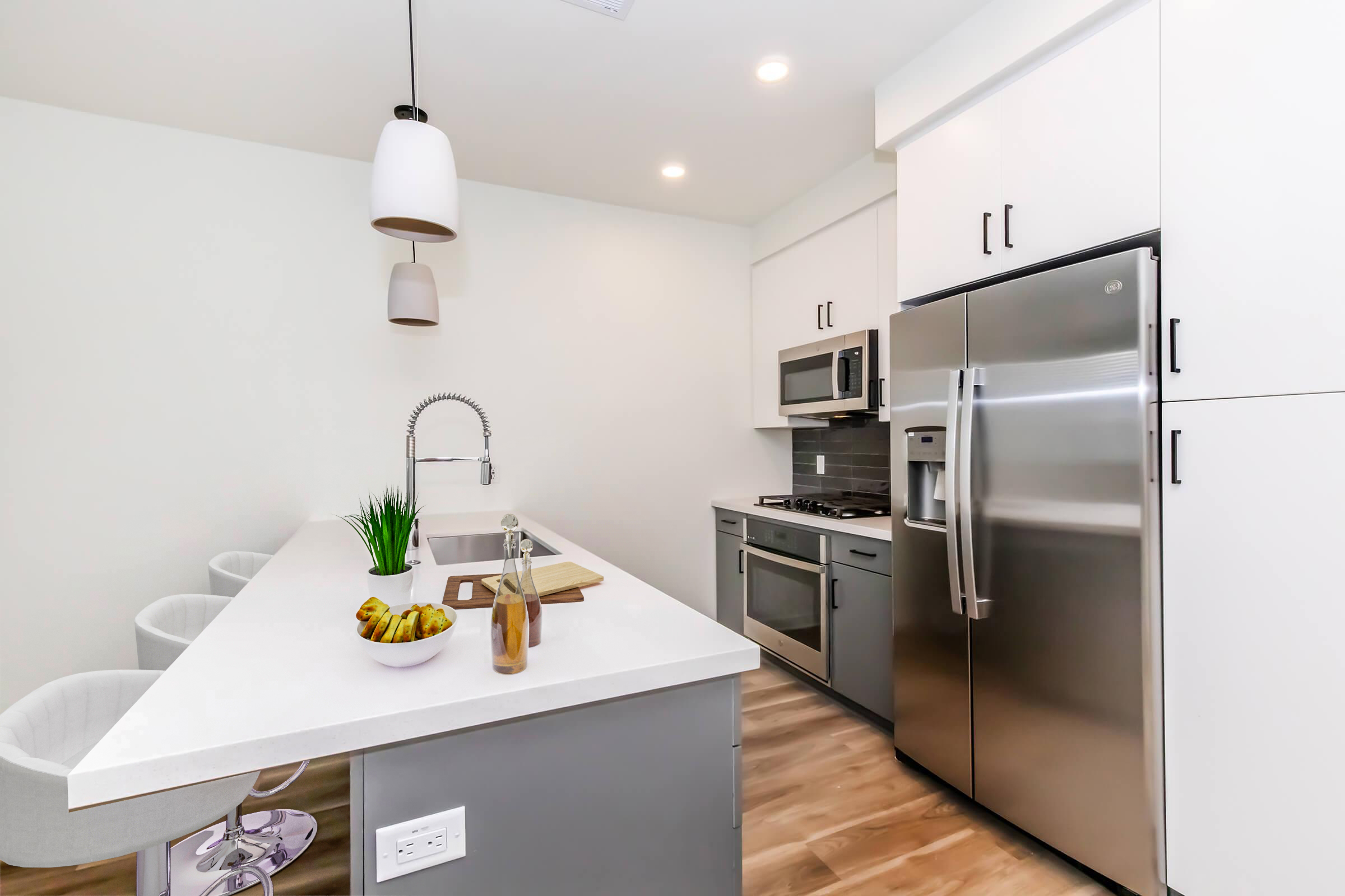 Modern kitchen featuring a sleek design with white cabinetry and stainless steel appliances. An island with stools, a cutting board, and a fruit bowl is highlighted, along with a faucet and overhead pendant lights. The space has warm wood flooring and neutral-colored walls, creating a clean and inviting atmosphere.
