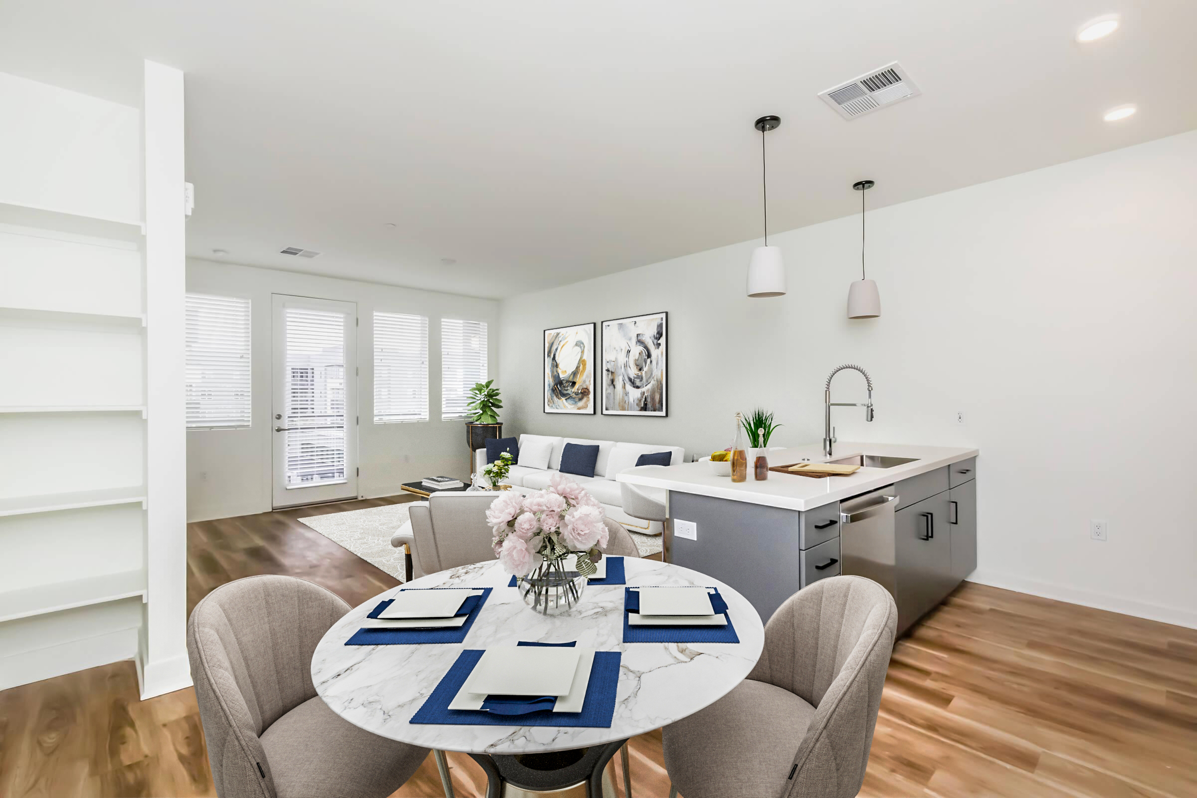A modern living space featuring a circular marble dining table set with plates and a floral centerpiece. In the background, a bright and airy lounge area with a light sofa, art on the walls, and large windows. A sleek kitchen island is partially in view, enhancing the contemporary feel of the room.