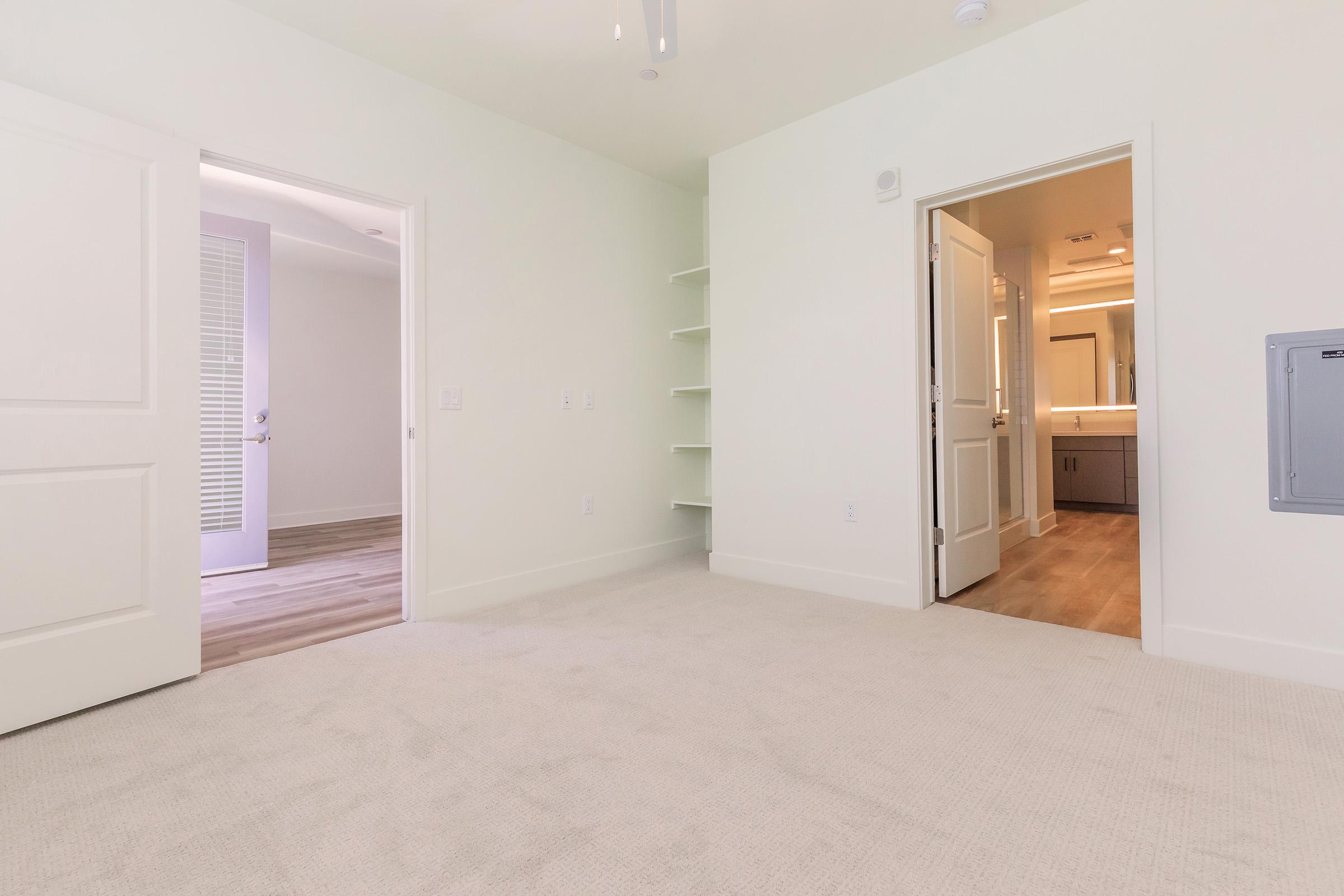 A well-lit, empty room with pale green walls and light beige carpeting. There are two open doors: one leads to a bathroom with a modern vanity and lighting, while the other opens to a hallway or another room. A shelf is mounted on the wall, adding storage options to the space.