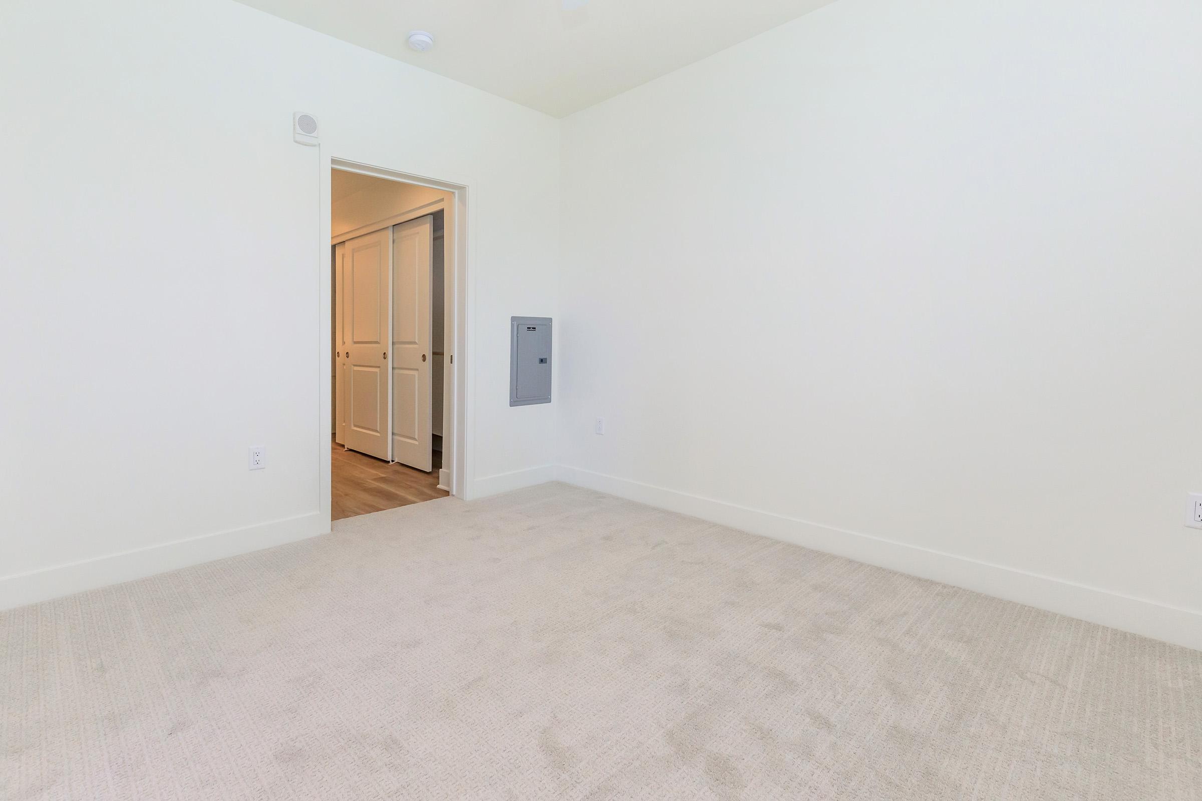 Empty room with light-colored walls and beige carpet. A doorway leads to another area, and there is a wall-mounted mailbox on the right. The space is well-lit and minimalist, suggesting a modern design.
