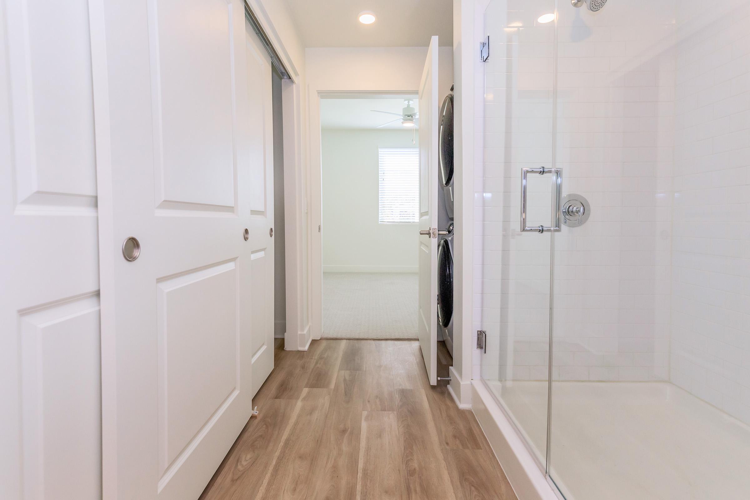 A modern laundry room with sliding doors, featuring a walk-in shower with transparent glass doors. The floor is made of light-colored wood, and there's a ceiling fan visible in the adjacent room. Natural light streams in through a window, creating a bright and airy atmosphere.