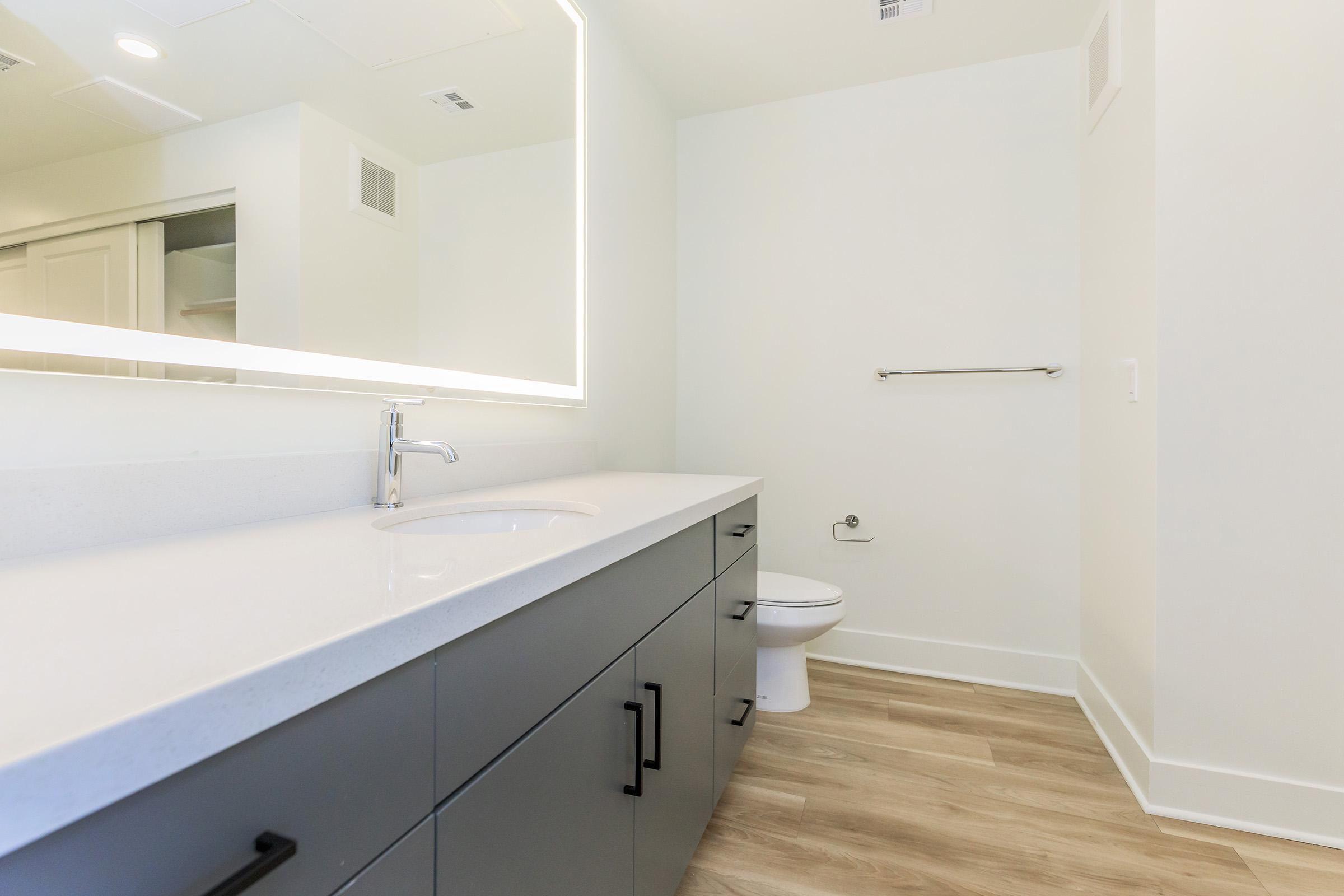 A modern bathroom featuring a large, sleek vanity with a white countertop and a circular sink. A backlit mirror is mounted above the vanity. The space has light-colored walls and wooden flooring, with a toilet positioned against the wall. There’s also a towel bar installed on the wall.