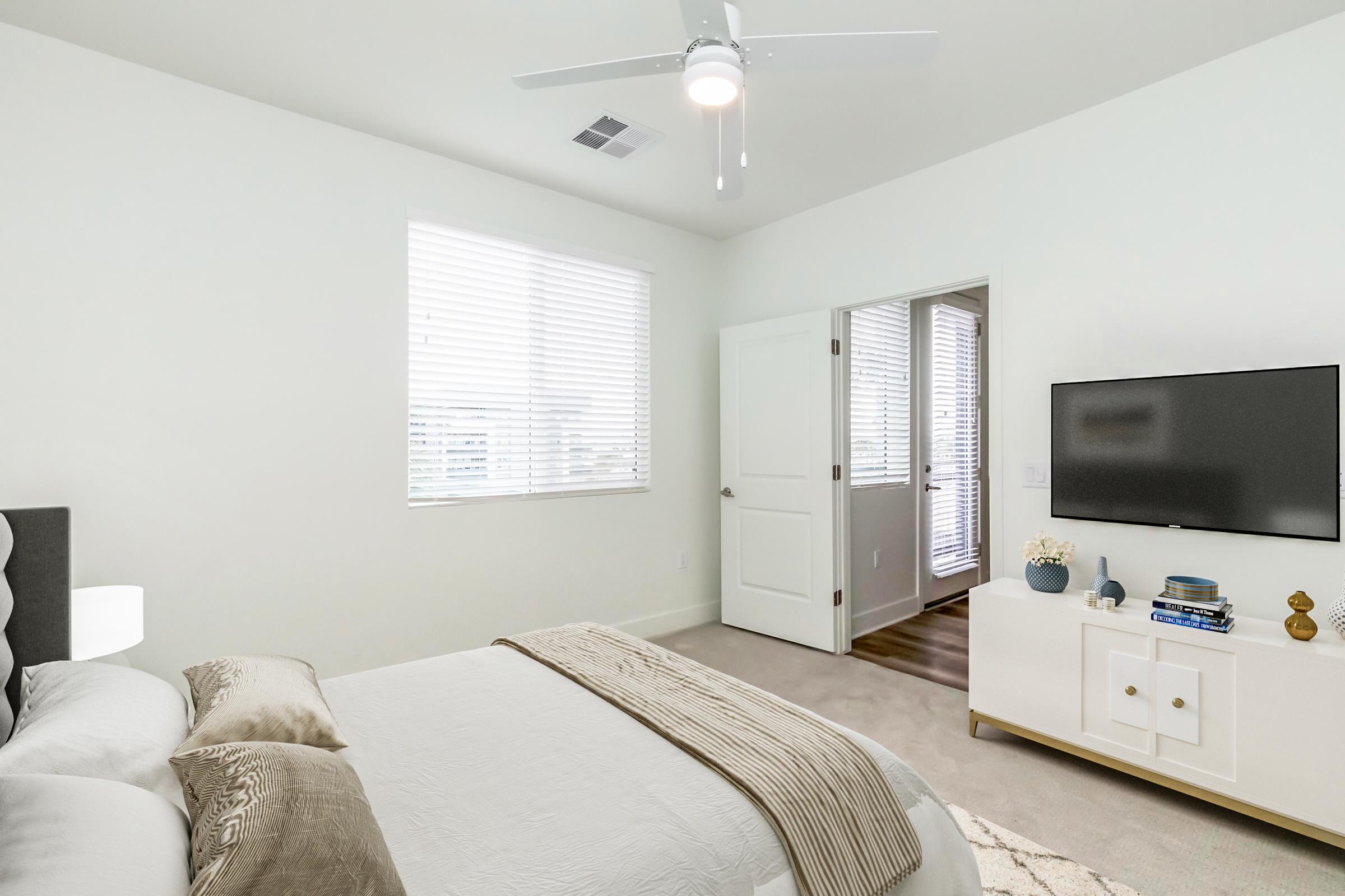 A modern bedroom featuring a neatly made bed with light bedding and decorative pillows. A ceiling fan is overhead, and the room is brightly lit by natural light coming through the window with white blinds. There’s a wall-mounted TV across from the bed and a stylish white cabinet beneath it, creating a minimalist aesthetic.