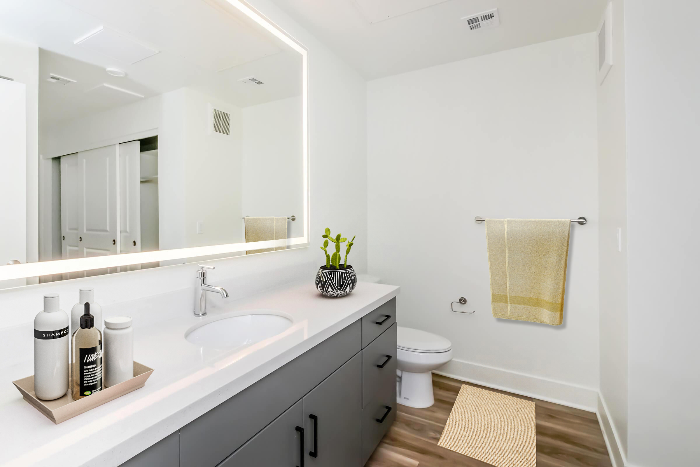 A modern bathroom featuring a sleek vanity with a round sink, a backlit mirror, and a decorative plant. The space includes a white toilet, a hanging towel in a soft yellow, and a small area rug on the wood-like floor. White walls and light cabinetry create a clean, fresh look.