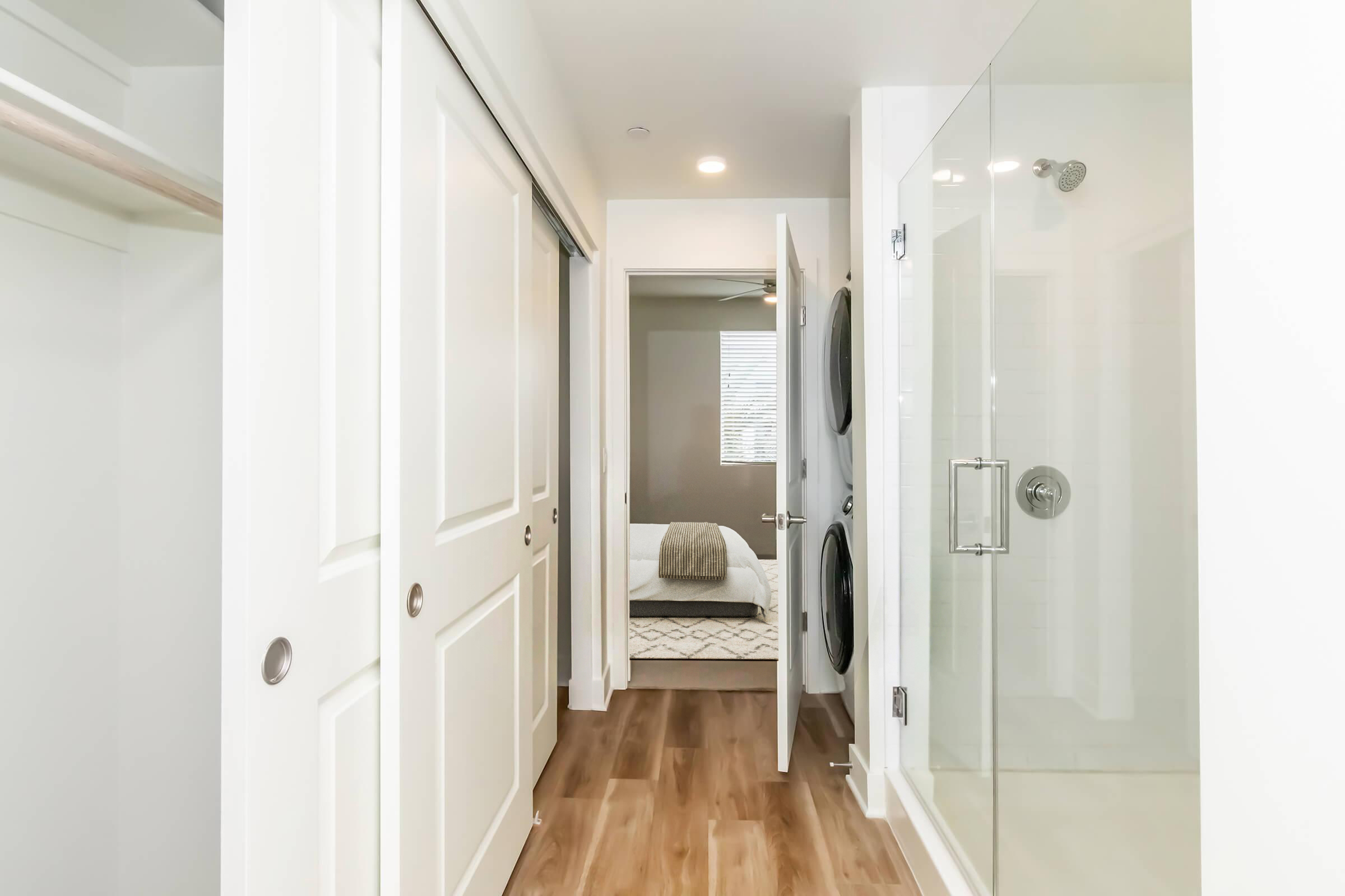 A modern hallway leading to a bathroom and bedroom. The hallway features white doors and a light wooden floor. On the right, there is a glass shower enclosure, and on the left, a laundry setup with a stacked washer and dryer. The bedroom visible in the distance has a bed with neutral bedding.