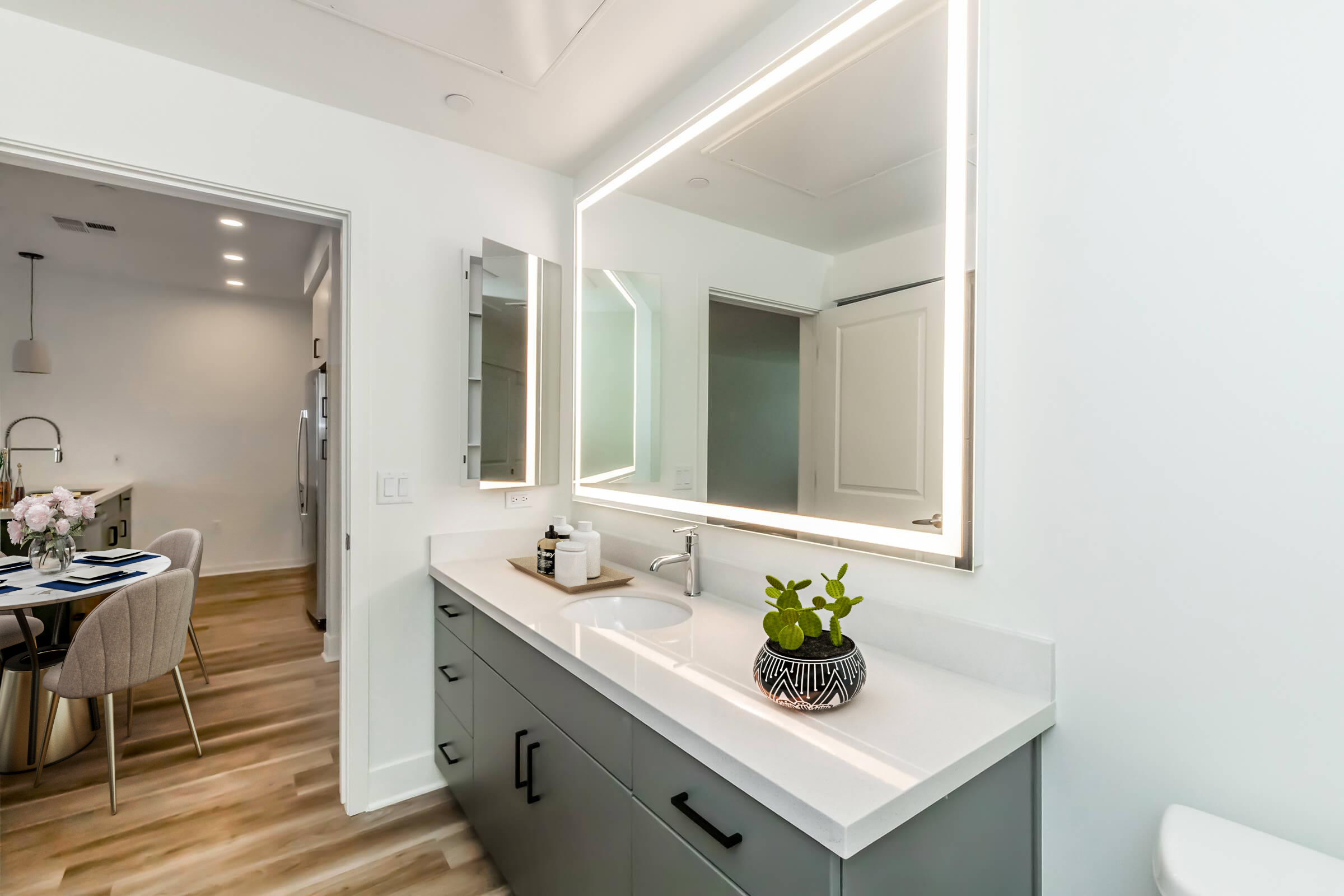 Modern bathroom featuring a sleek, dual-sink vanity with a white countertop and dark gray cabinetry. Above the sinks, there's a large backlit mirror. A small potted plant sits on the counter. In the background, a glimpse of a dining area can be seen, with a round table and chairs. Bright, neutral color scheme throughout.