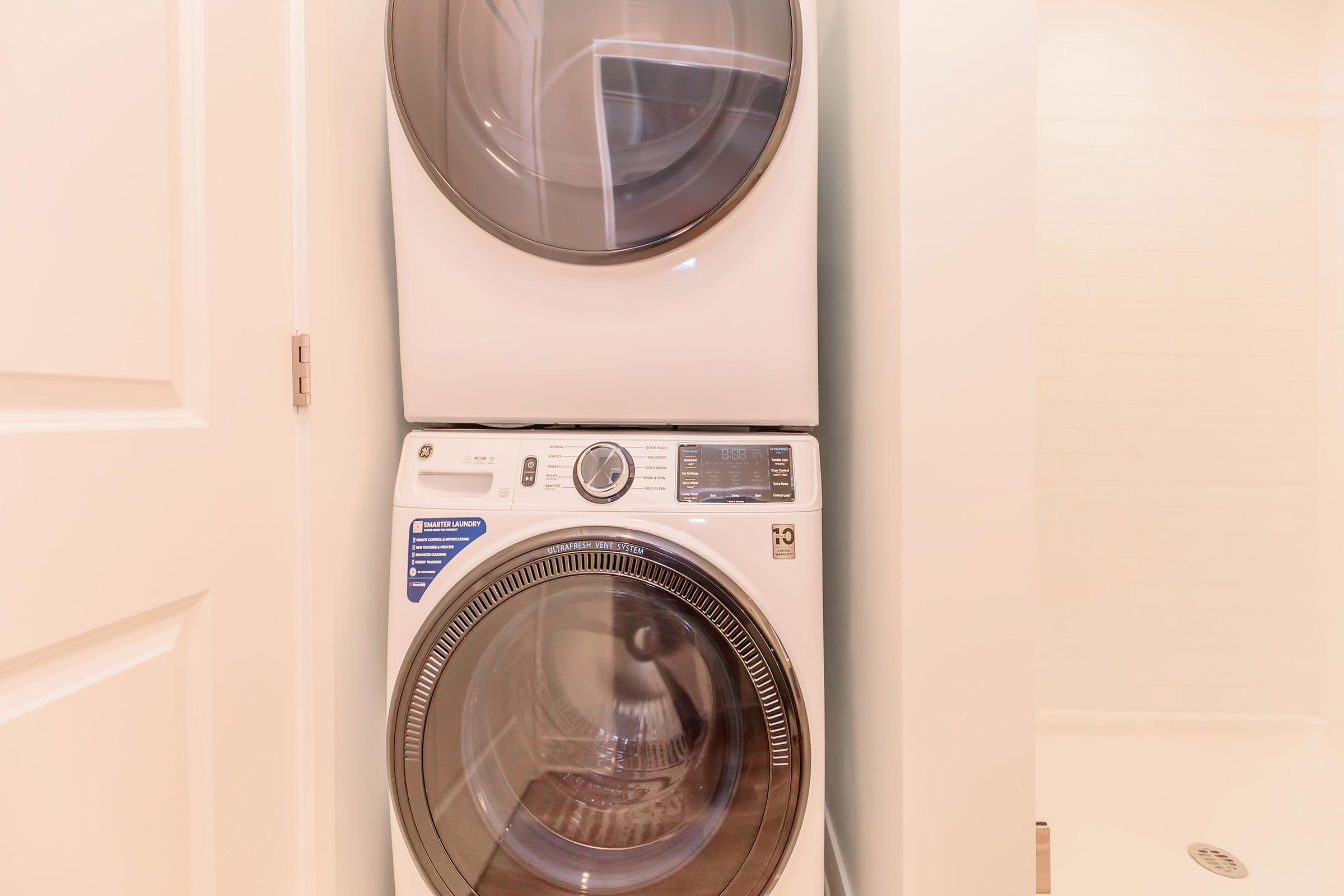 Stacked washer and dryer in a narrow laundry space. The washer is at the bottom with a front-loading design, while the dryer is above it. The appliances are modern and white, positioned between a door and a shower area, with light-colored walls in the background.