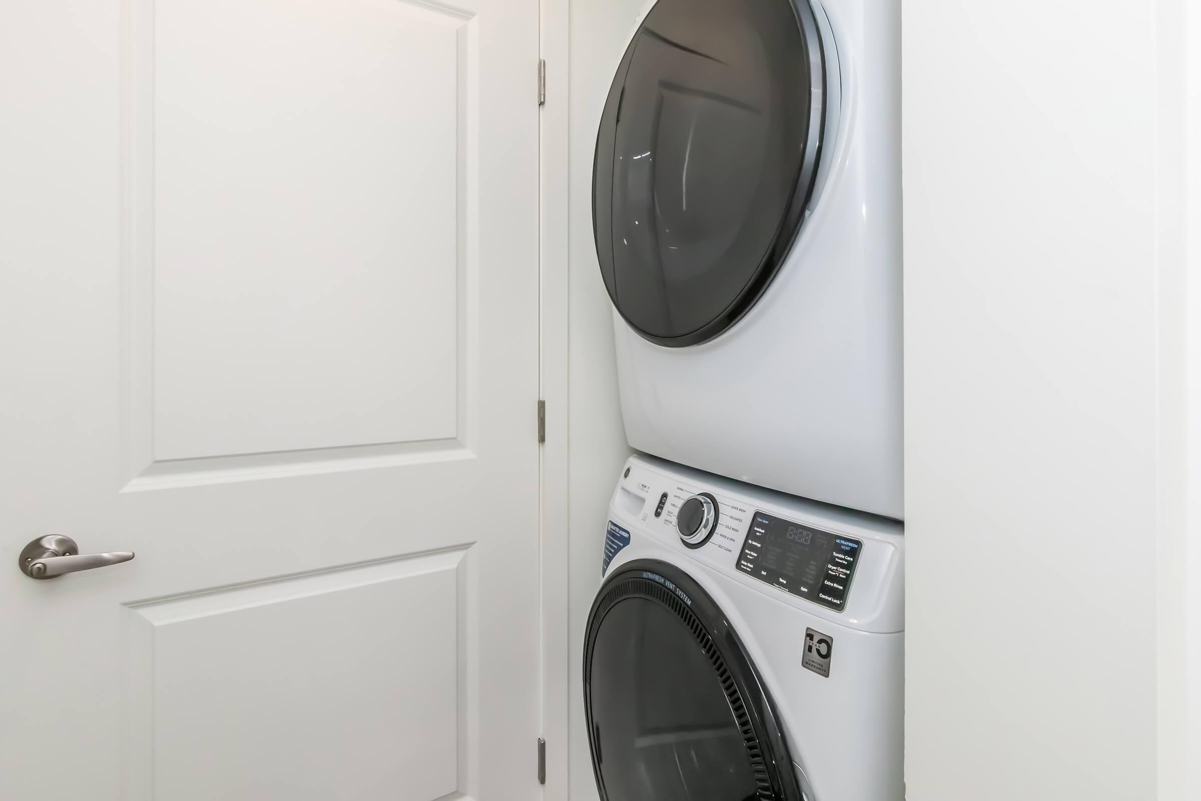 A stacked washer and dryer set in a narrow laundry space, positioned near a white door. The appliances have a modern design with a digital control panel visible. Natural light may be filtering in, illuminating the clean, minimalistic laundry area.