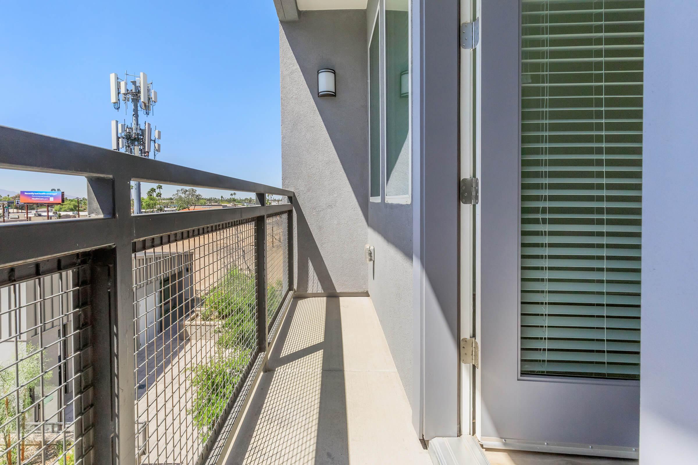 A view of a balcony with a modern design, featuring a metal railing and sliding glass door. The space is well-lit with clear blue skies and a cellular tower visible in the background, along with some greenery below. The scene suggests a contemporary urban environment.