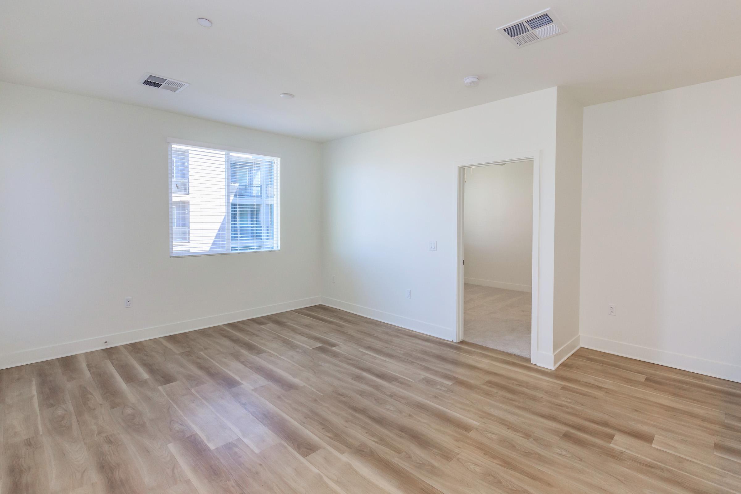 A spacious, empty room with light-colored walls and hardwood flooring. There is a window allowing natural light in, and an open doorway leading to another room. The space has a modern feel with minimal decor, ideal for various furnishings or uses.