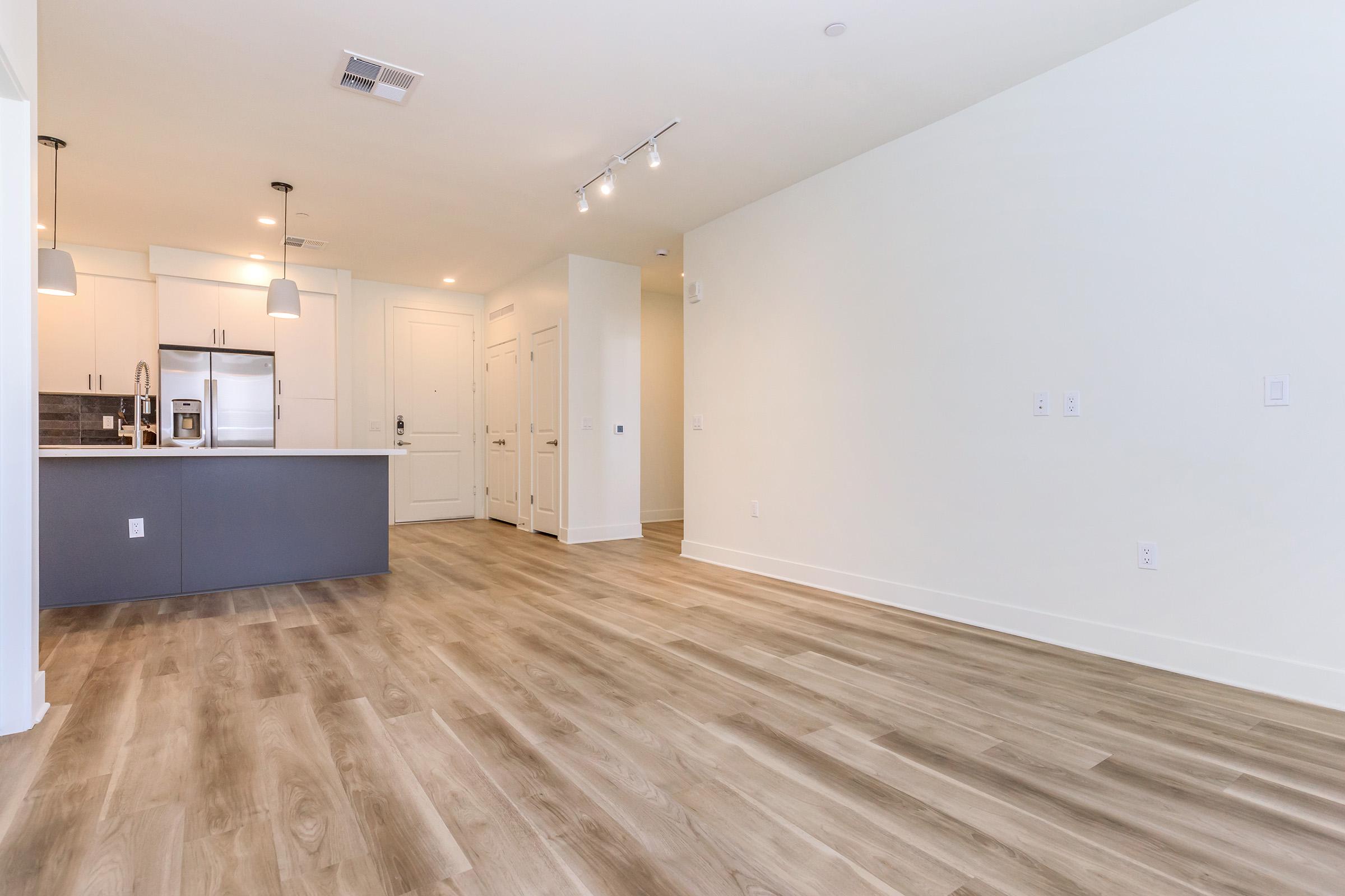 A spacious, modern living area with light-colored walls and a wooden floor. The kitchen is in the background, featuring sleek cabinets and stainless steel appliances. Recessed lighting illuminates the space, which is designed for an open floor plan. Multiple doors lead to additional rooms or storage.