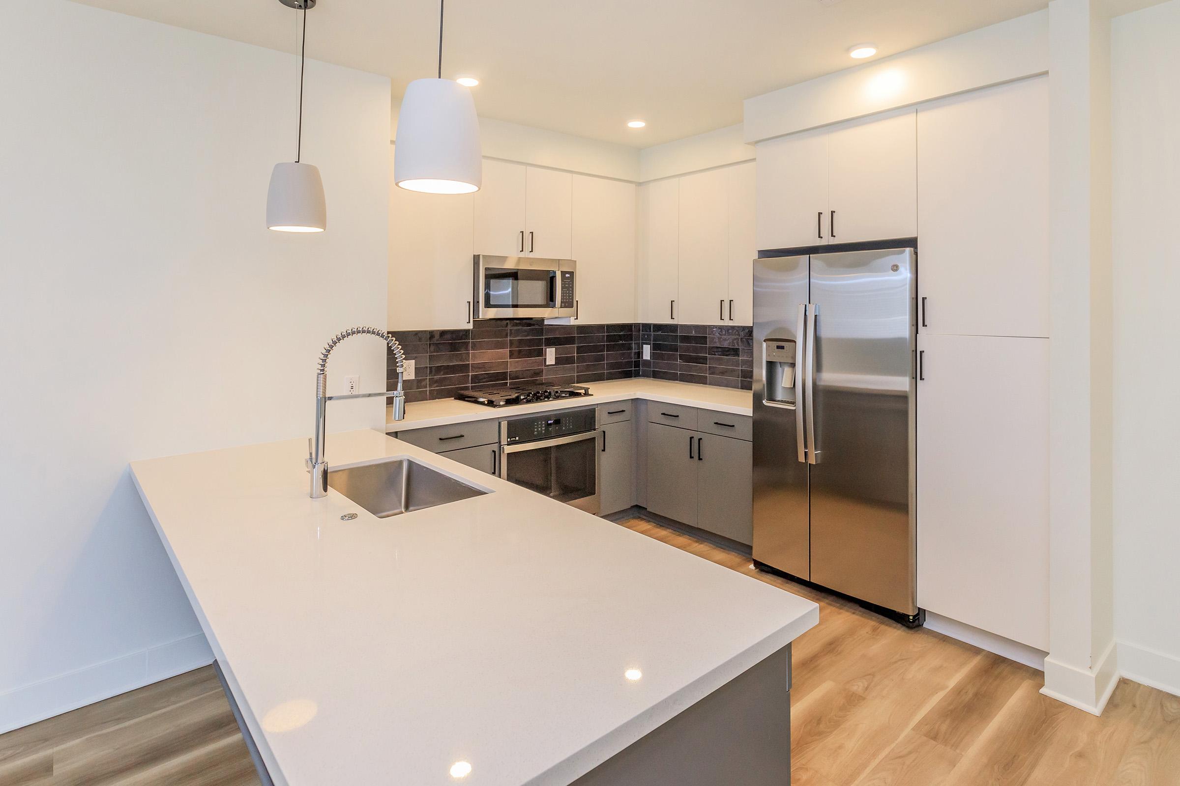 Modern kitchen featuring a central island with a sink, stainless steel appliances, including a microwave and refrigerator, gray cabinetry, and white countertops. The space is illuminated by pendant lights and has a sleek, contemporary design with a wood-like flooring.