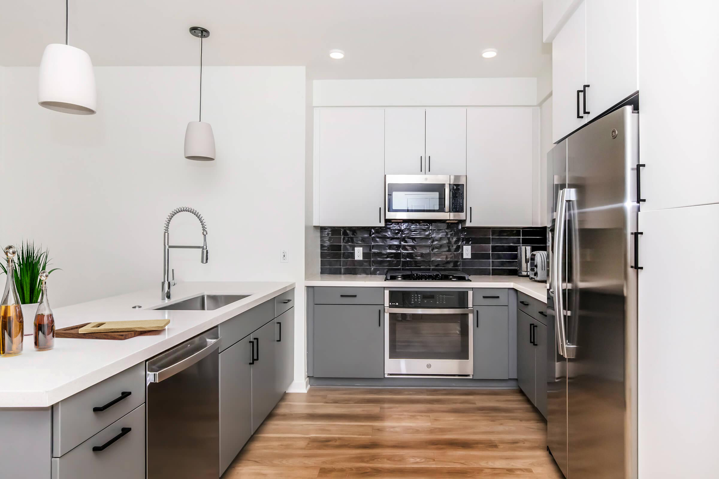 Modern kitchen with gray cabinets and stainless steel appliances, featuring a large sink, a built-in microwave, and a sleek refrigerator. White countertops and pendant lighting create a bright, clean aesthetic, while a decorative plant adds a touch of warmth. Wood flooring completes the contemporary look.