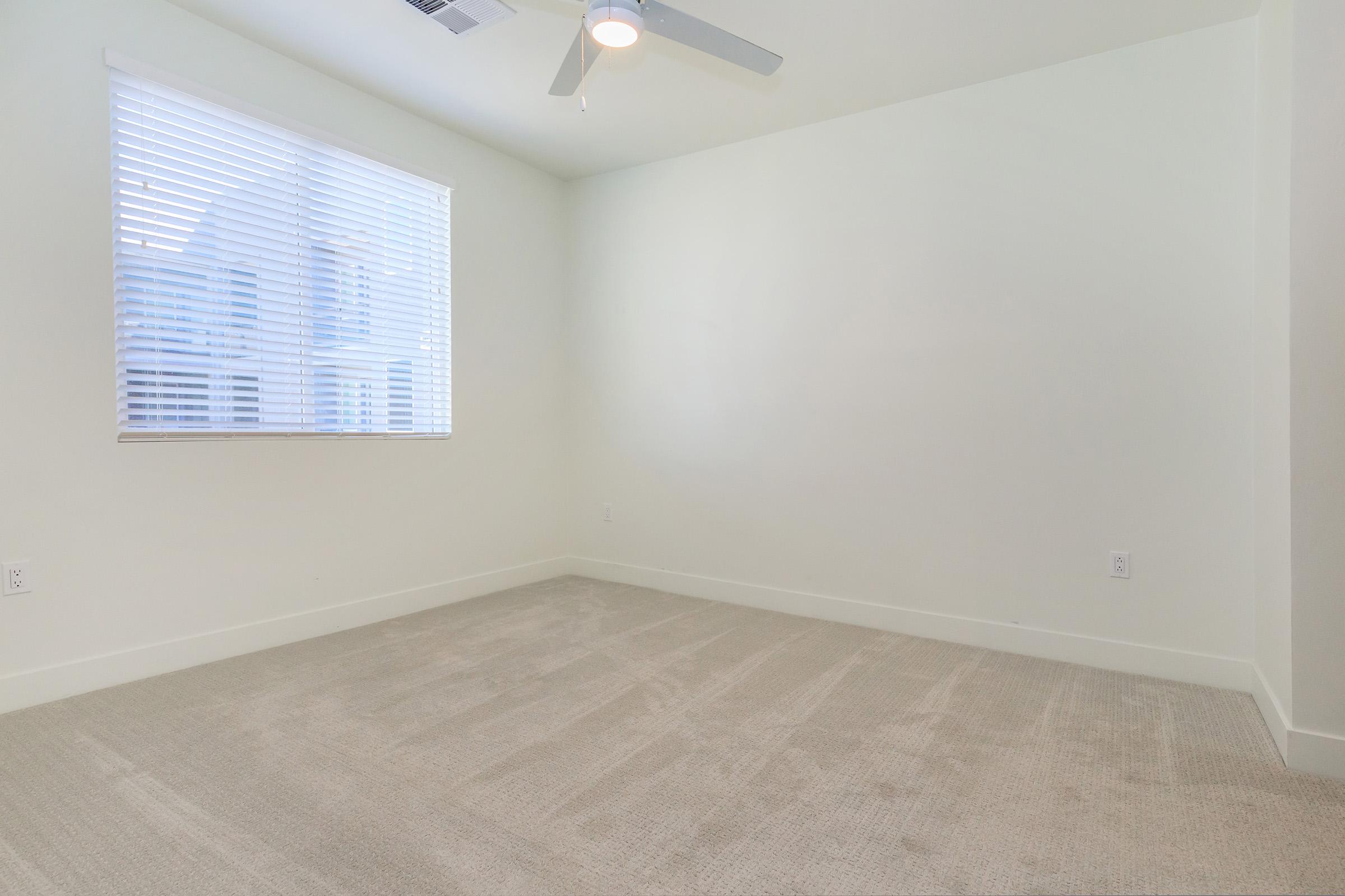 A spacious, empty room with light-colored walls and a beige carpet. A window with horizontal blinds allows natural light to enter the space, and a ceiling fan is visible. The room has a clean and minimalist appearance, suitable for various purposes.