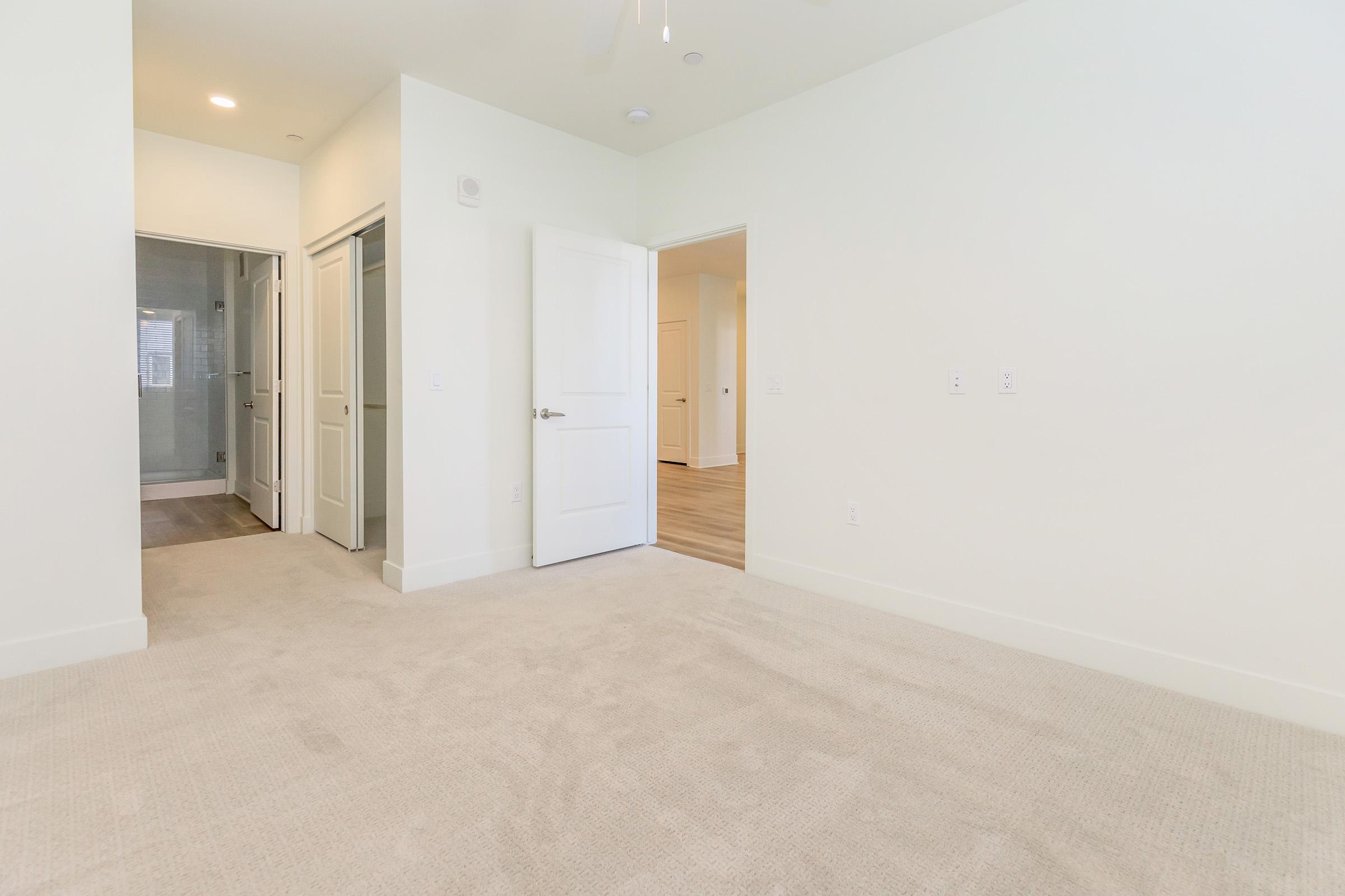 A spacious, empty room with light-colored walls and plush beige carpet. Two doorways lead to adjacent areas, one on the left and another in the background. Soft natural light enters through unseen windows, creating a bright and inviting atmosphere.