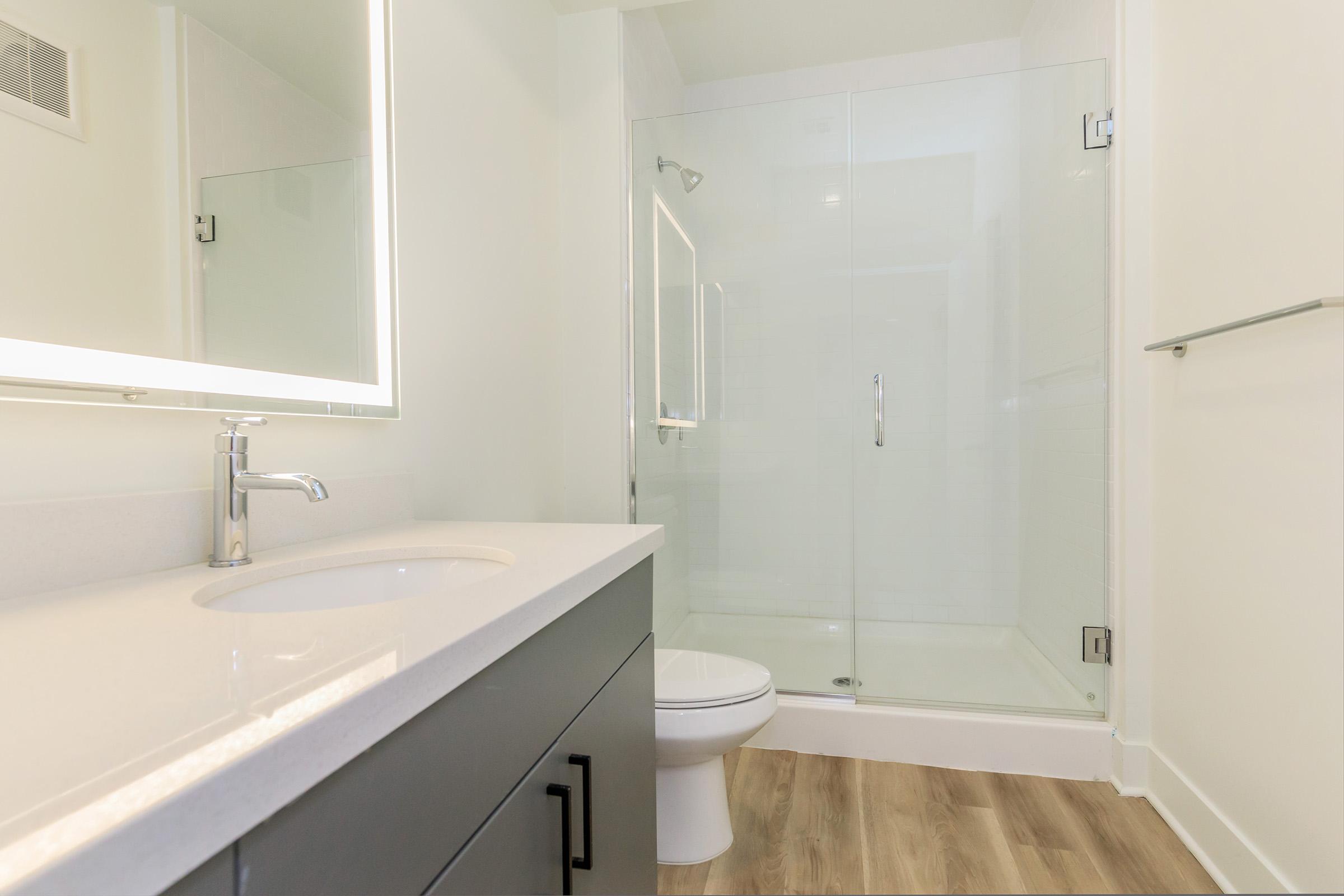 A modern bathroom featuring a sleek gray vanity with a white countertop and sink, a large illuminated mirror, and a glass-enclosed shower. The space has neutral-colored walls and wood-like flooring, creating a bright and contemporary atmosphere.