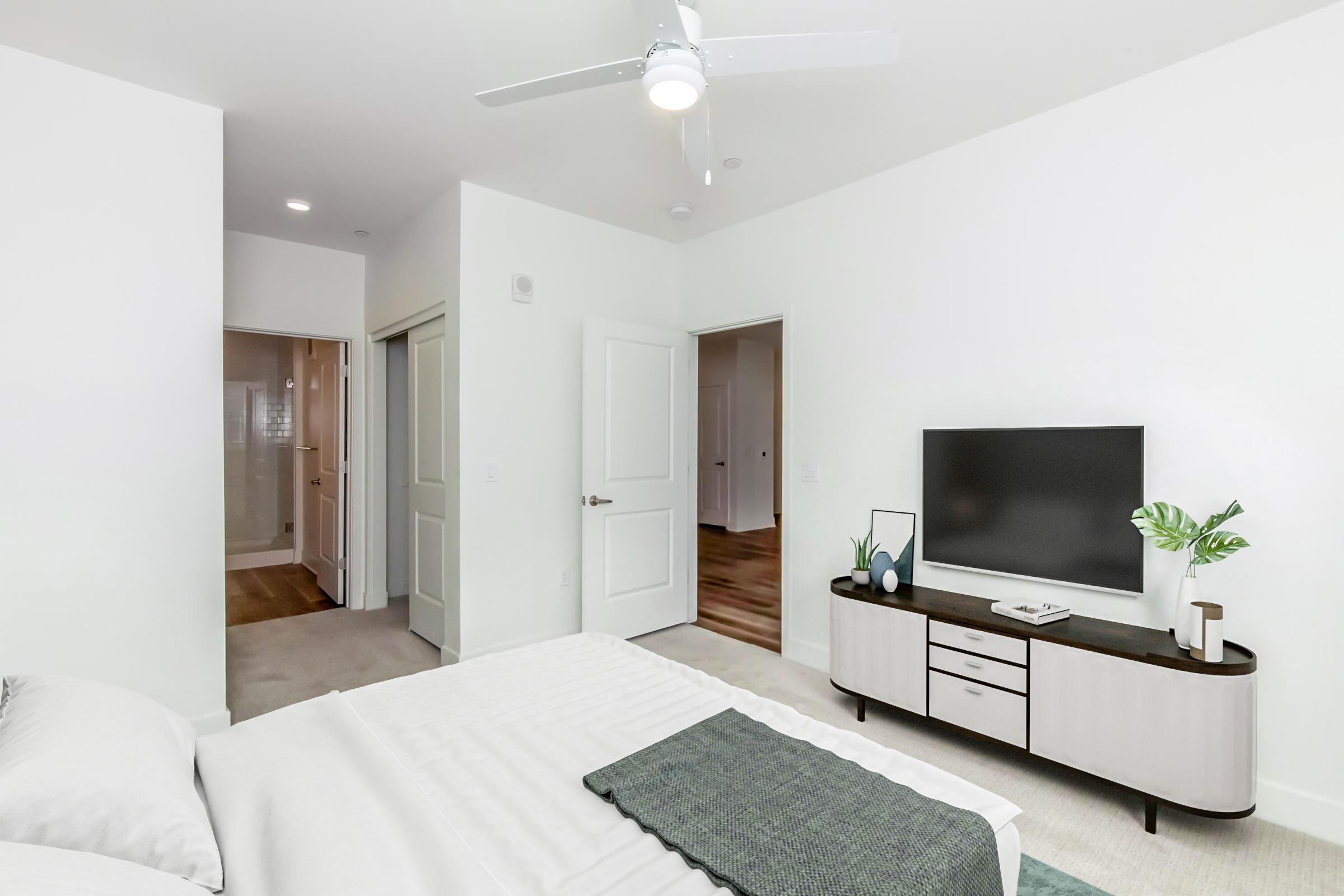 Modern bedroom with a cozy bed covered in a light comforter, a sleek dresser with a TV on top, and a decorative plant. A ceiling fan is visible, and there are white walls and a doorway leading to a bathroom. Natural light brightens the space, creating a serene and inviting atmosphere.