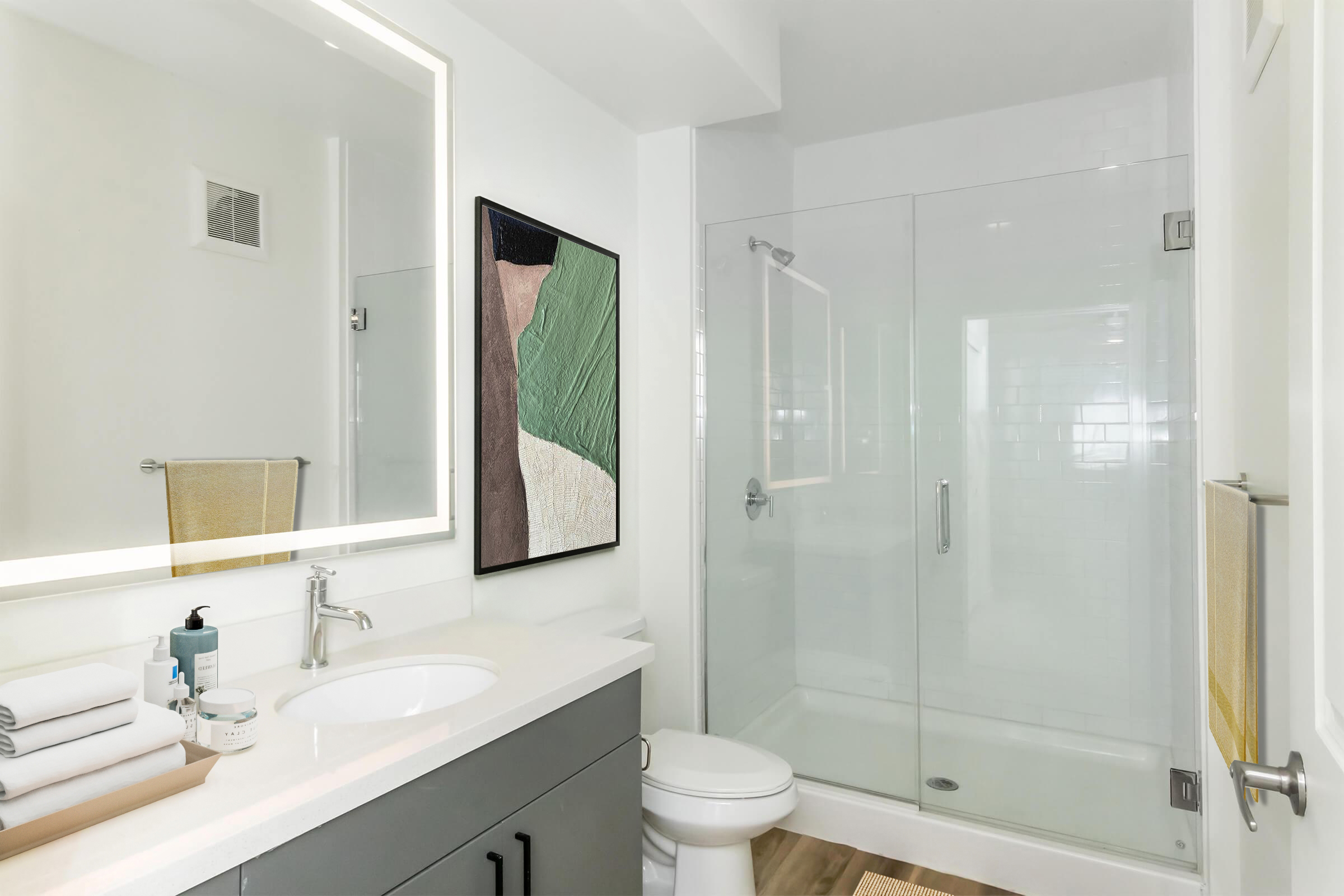 A modern bathroom featuring a glass shower enclosure and a white countertop with a sink. The space is illuminated by a backlit mirror, and artwork is on the wall. Towels are neatly arranged on the counter, and there are wooden accents on the floor, creating a clean and contemporary look.