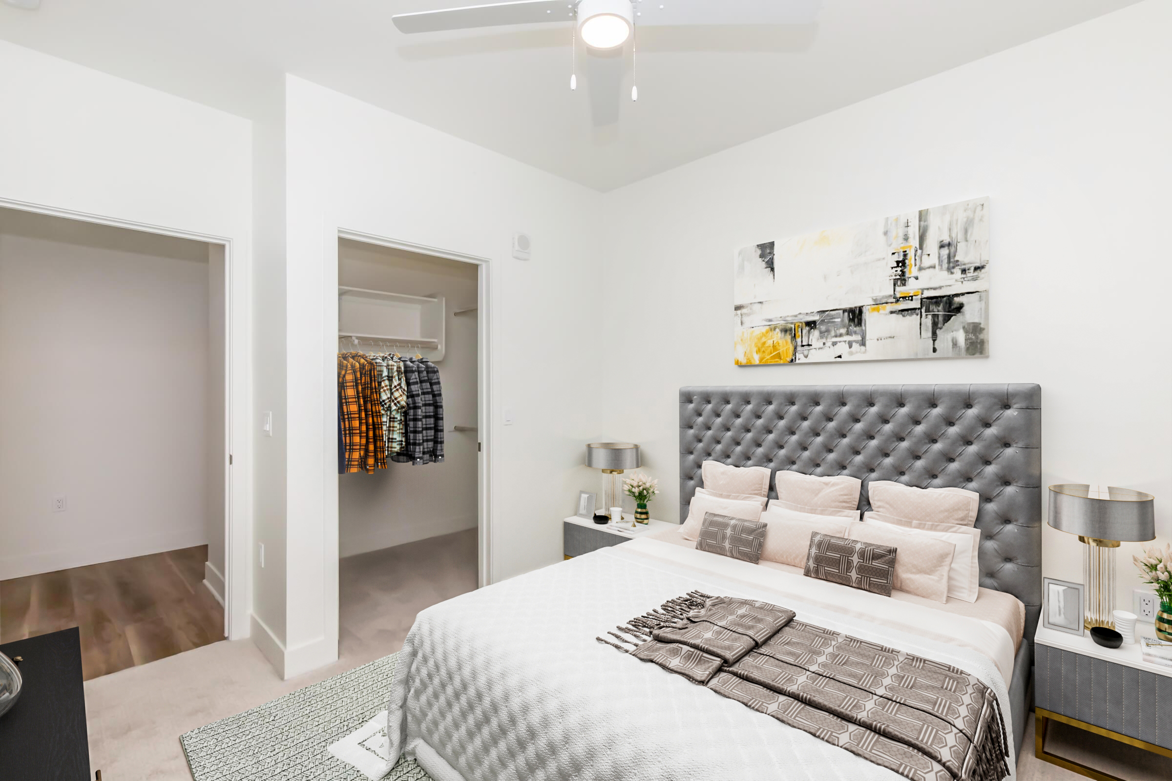 A modern bedroom featuring a gray upholstered bed with decorative pillows, a bedside table with lamps, and a large abstract painting above the headboard. There's a closet with neatly hung plaid shirts visible in the background, and the room has light-colored walls and a ceiling fan.