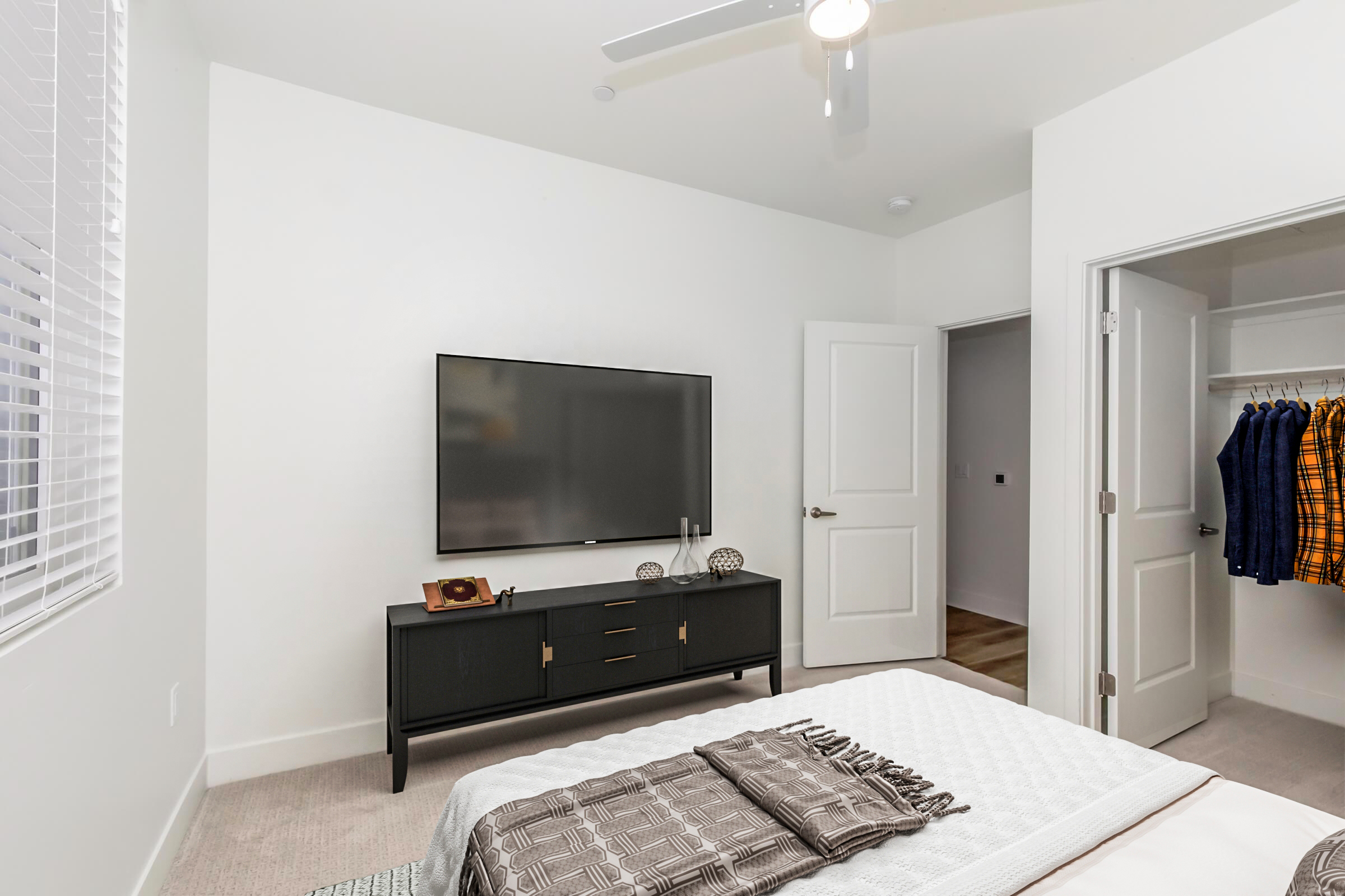 A modern bedroom featuring a white wall, light-colored carpet, a black TV stand with a television, and decorative items on top. A cozy bed with a patterned throw blanket is in the foreground, while a closet with hanging clothes is visible through an open door. A ceiling fan is mounted above.