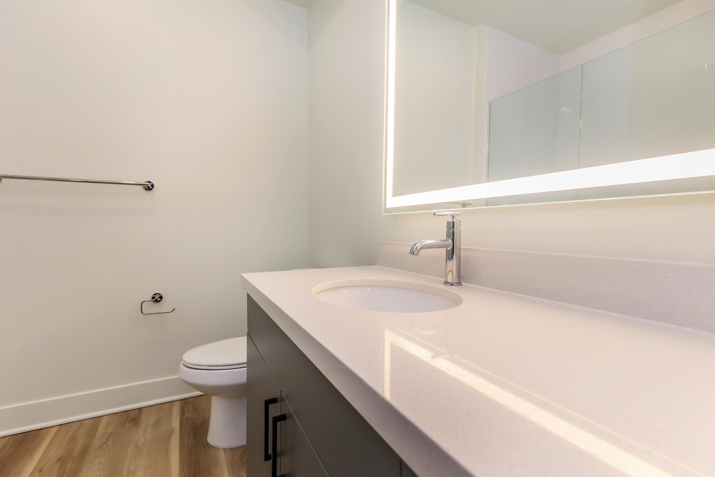 A modern bathroom featuring a sleek, light-colored vanity with a white countertop and sink. There is a large mirror with integrated lighting above the sink. The floor is wooden, and a toilet is visible in the background, along with a towel bar. The walls are painted a soft, neutral color.
