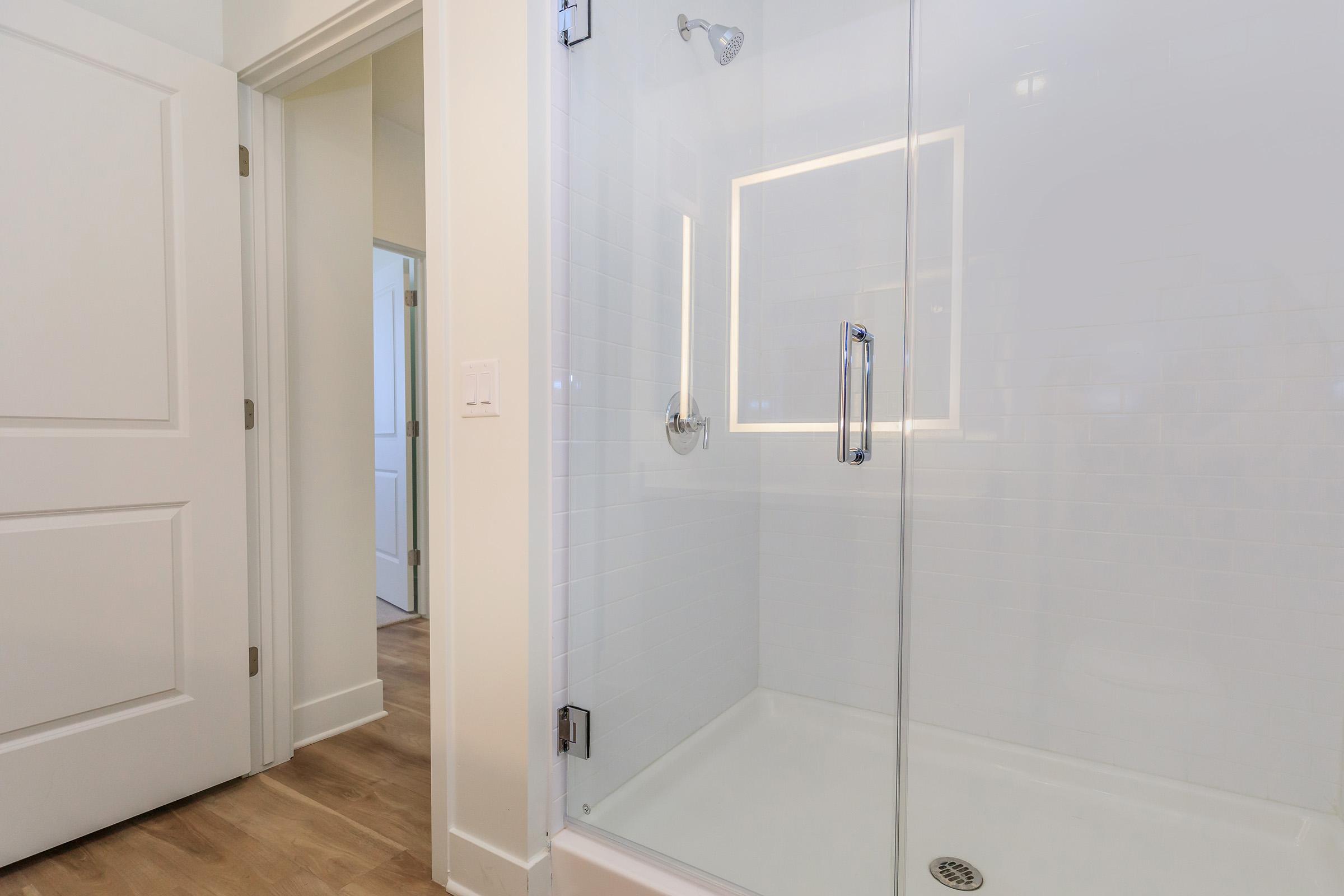 A modern bathroom featuring a glass shower enclosure with a sleek showerhead, white tiled walls, and a minimalist design. A glimpse of a door leading to another room is visible, showcasing a clean and contemporary interior. The flooring is a light wood finish, adding warmth to the space.