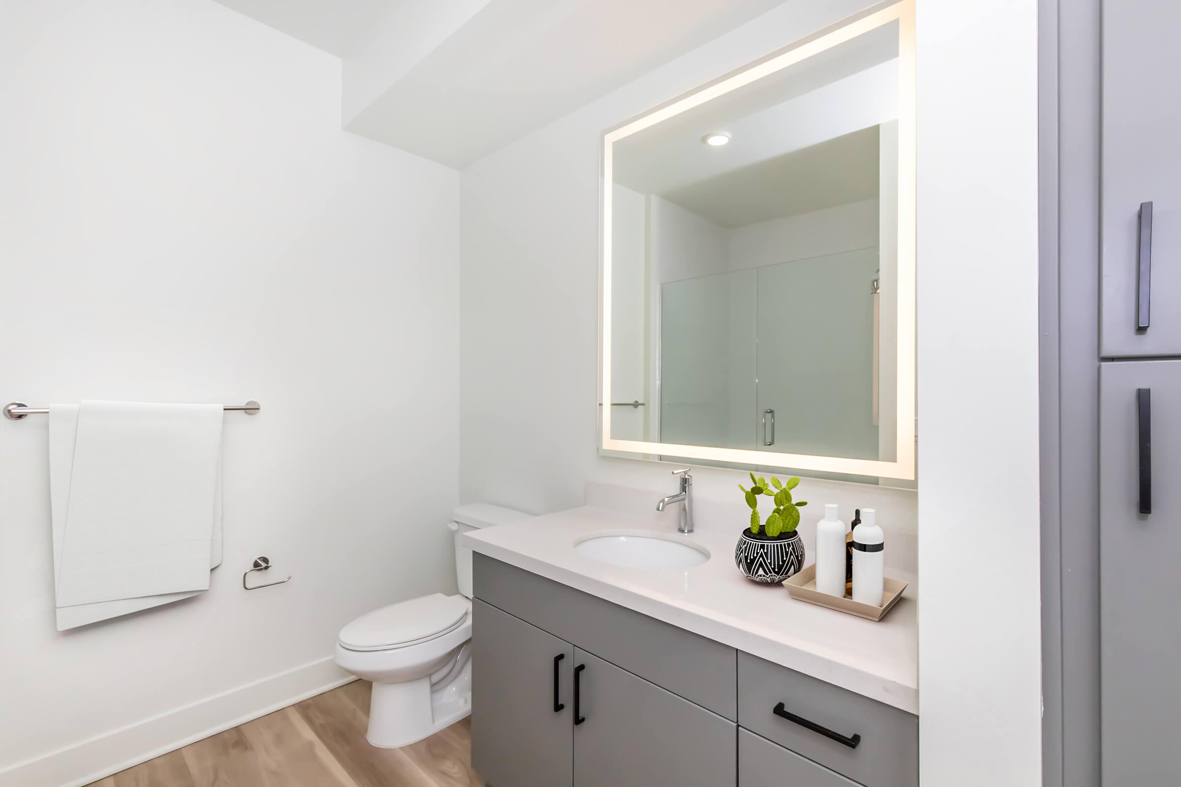 Modern bathroom featuring a sleek countertop with a sink, a large illuminated mirror, a towel rack with a white towel, and a minimalistic decor. The gray cabinetry blends with light walls and a wooden floor, while a small plant and toiletries adorn the countertop. A toilet is positioned in the corner.