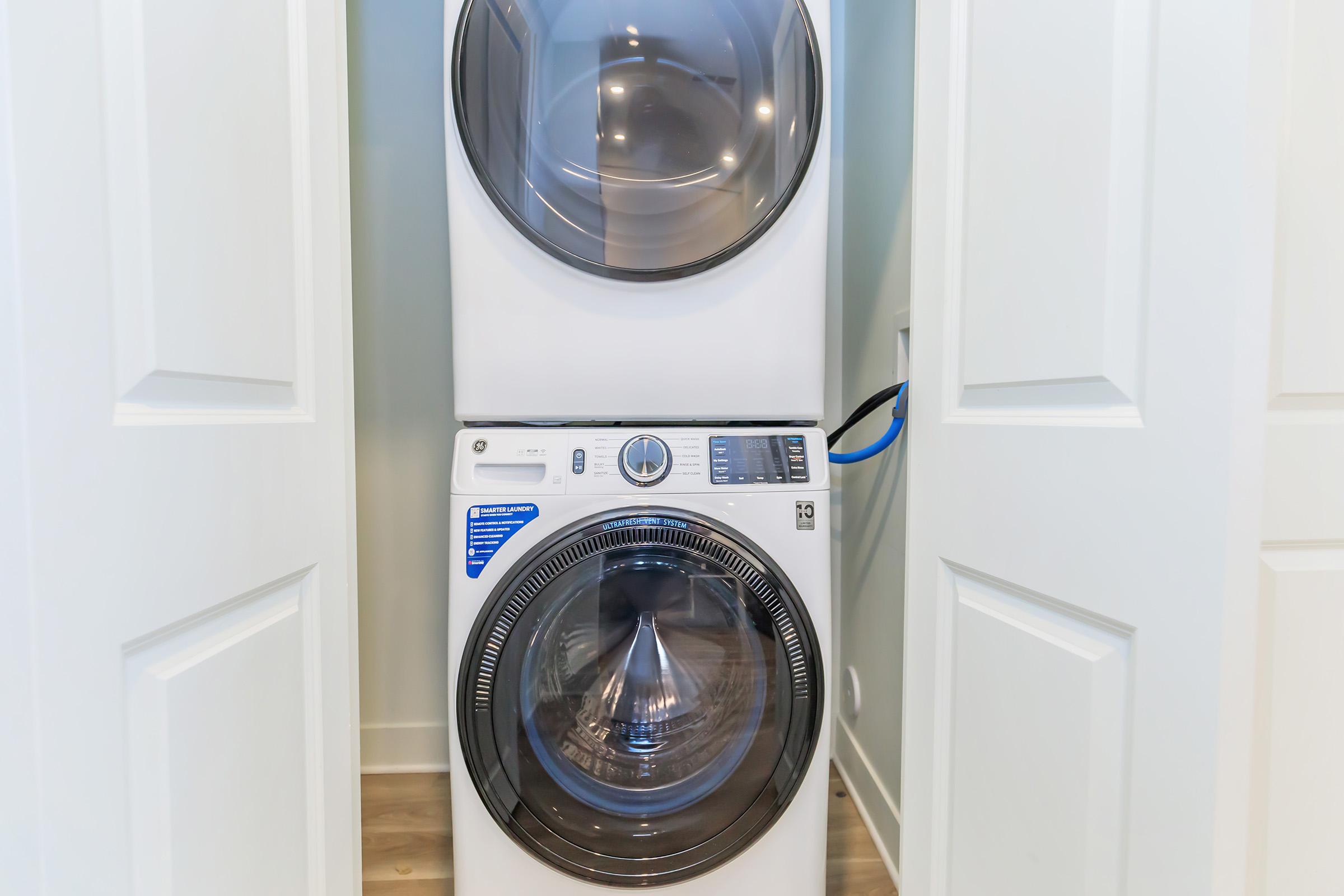 A stacked washer and dryer unit in a laundry space, positioned between white closet doors. The appliances are modern and display a clean, white exterior, with the dryer on top and the washer below. The space is well-lit with light-colored walls and wooden flooring.