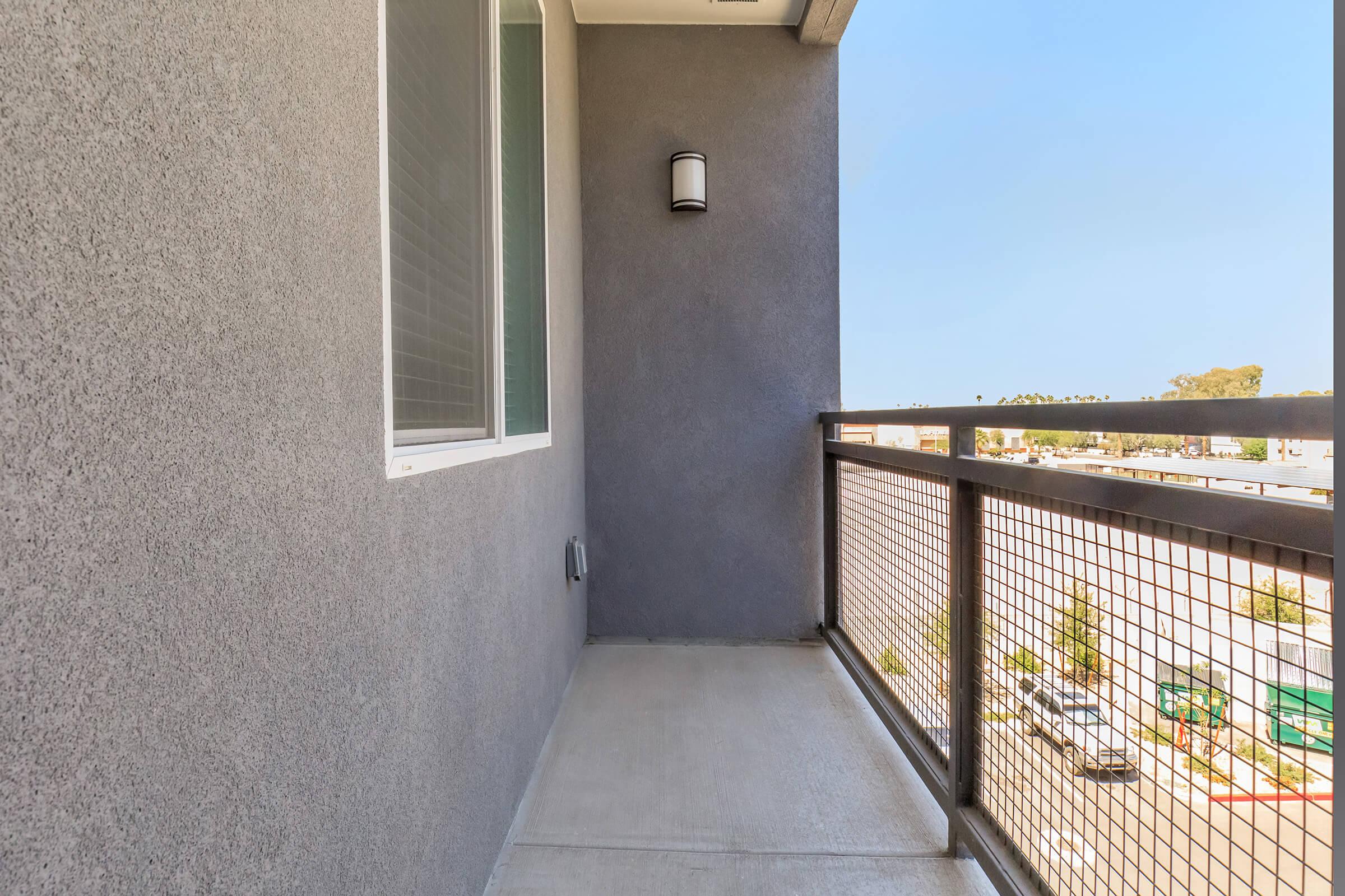 A concrete balcony with a gray wall and a light fixture on the side. There is a view of a parking lot and buildings in the distance. The floor is smooth and appears to be well-maintained, with a railing providing safety and a sense of openness. Clear blue sky is visible above.