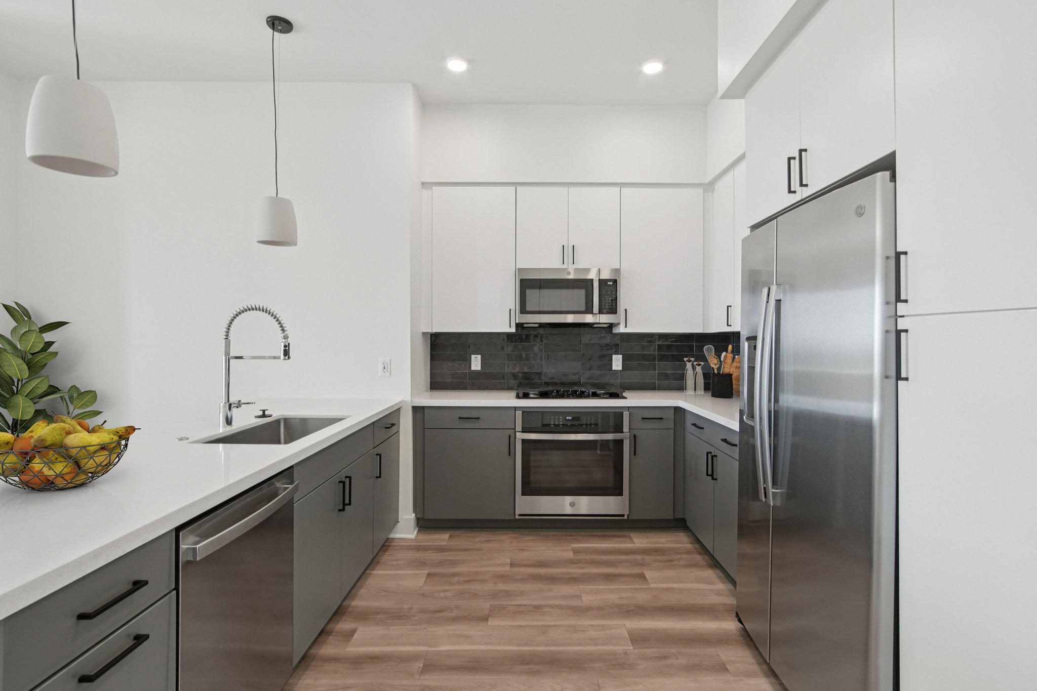 Modern kitchen featuring sleek gray cabinetry and stainless steel appliances. The layout includes a large sink, built-in microwave, and an oven. Natural light floods the space, highlighting a minimalist design. A fruit basket sits on the counter, adding a touch of color. Wood-style flooring complements the overall aesthetic.