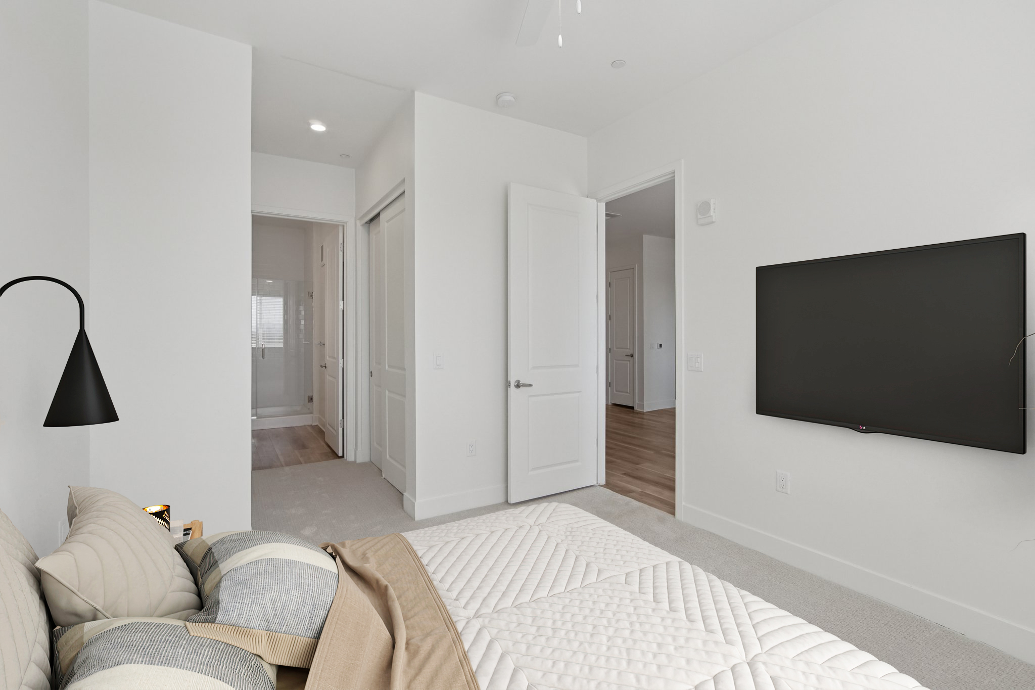 A modern bedroom featuring a neatly made bed with decorative pillows, a wall-mounted TV, and light-colored walls. The room has a doorway leading to a hallway and a bathroom visible in the background. A sleek floor lamp is positioned beside the bed, enhancing the minimalist decor.