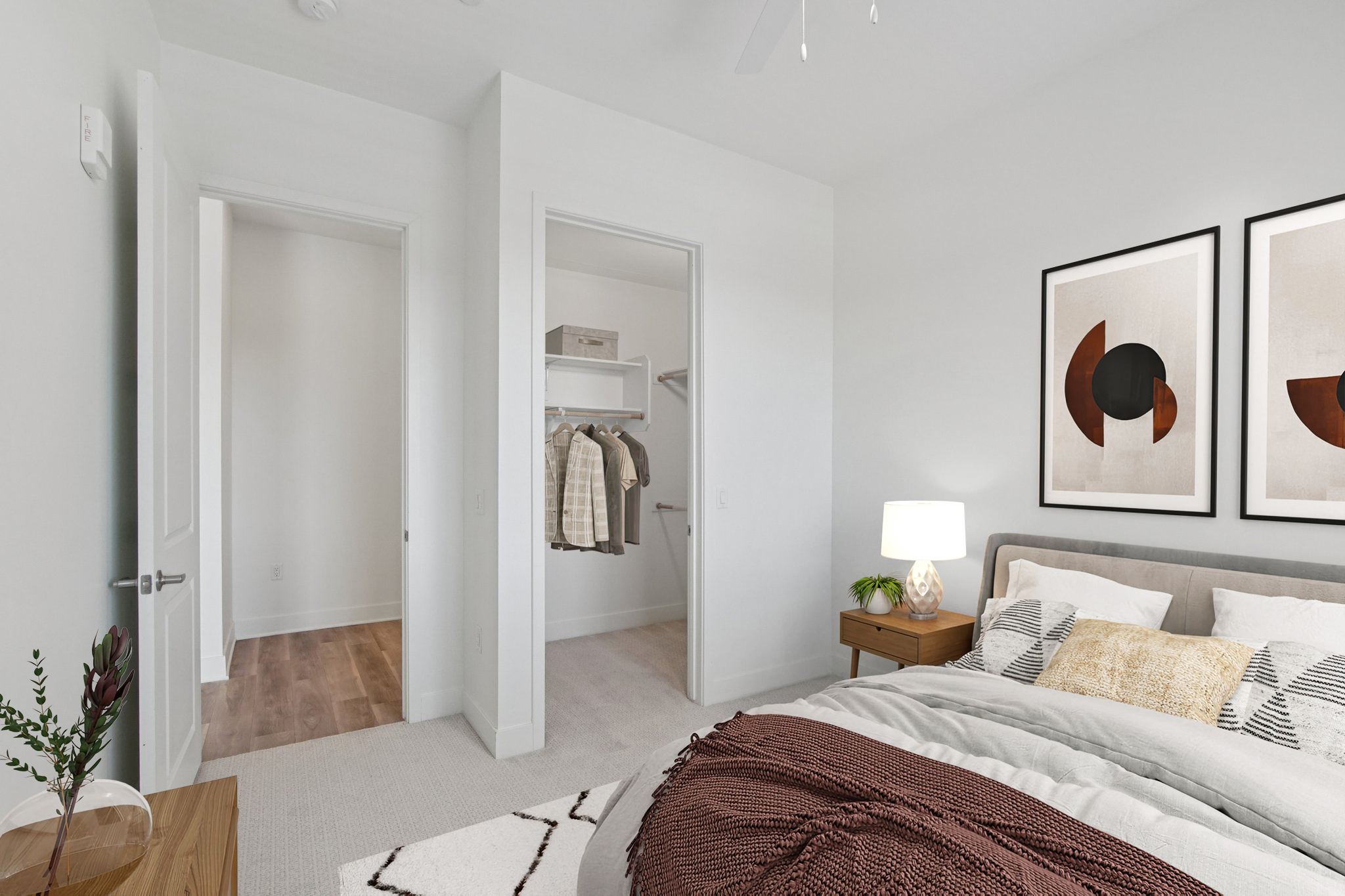 Cozy bedroom featuring a light-colored wall, a neatly made bed with decorative pillows and a patterned throw blanket. A bedside table with a lamp sits next to the bed. Doorways lead to a closet and another room. Natural light illuminates the space, creating a warm and inviting atmosphere.