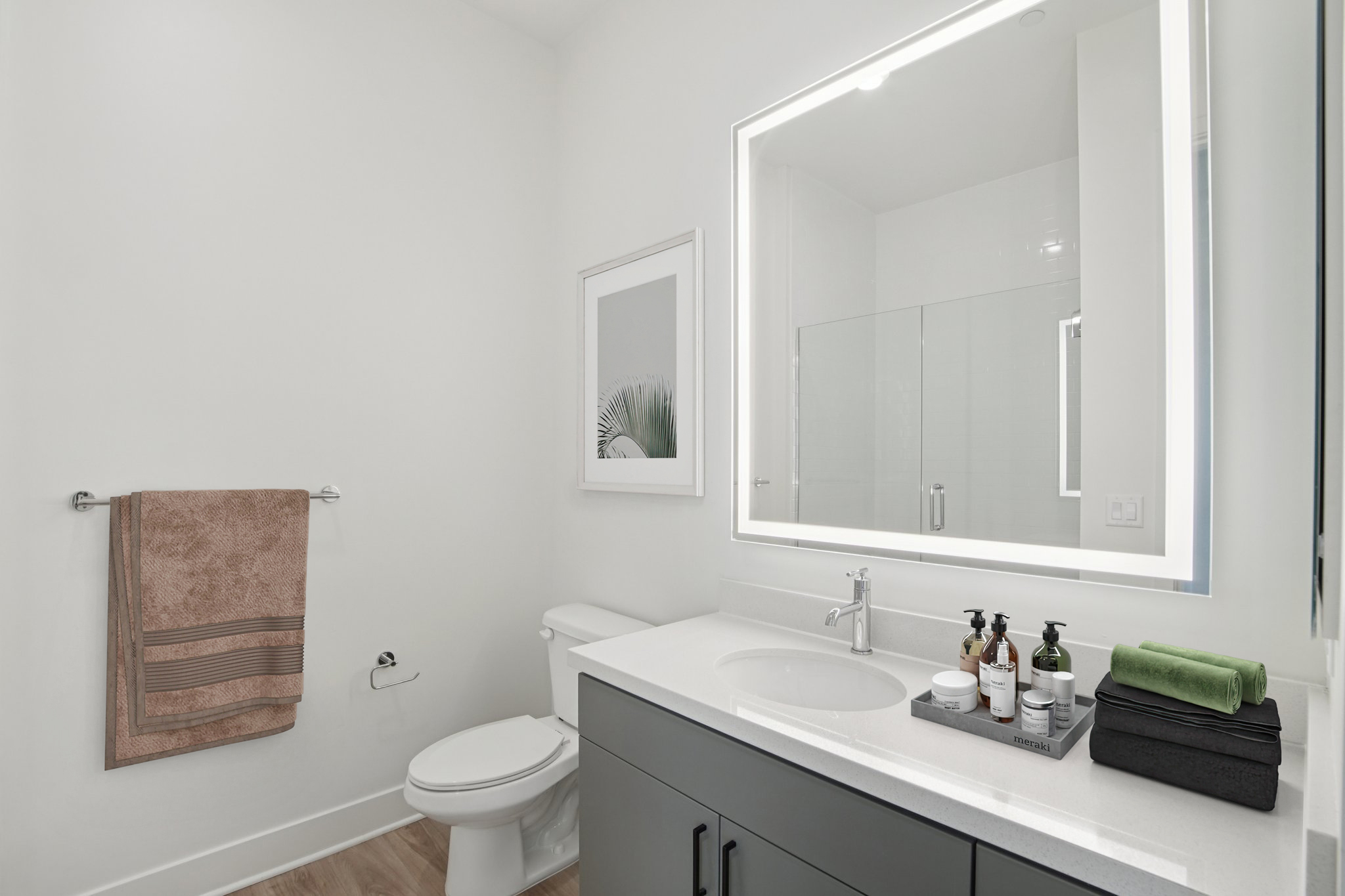 Modern bathroom featuring a toilet, a sleek vanity with a mirror and sink, and neatly arranged toiletries. A hand towel is hanging on a rack, and the floor has light wood-like tiles, creating a clean and stylish aesthetic.