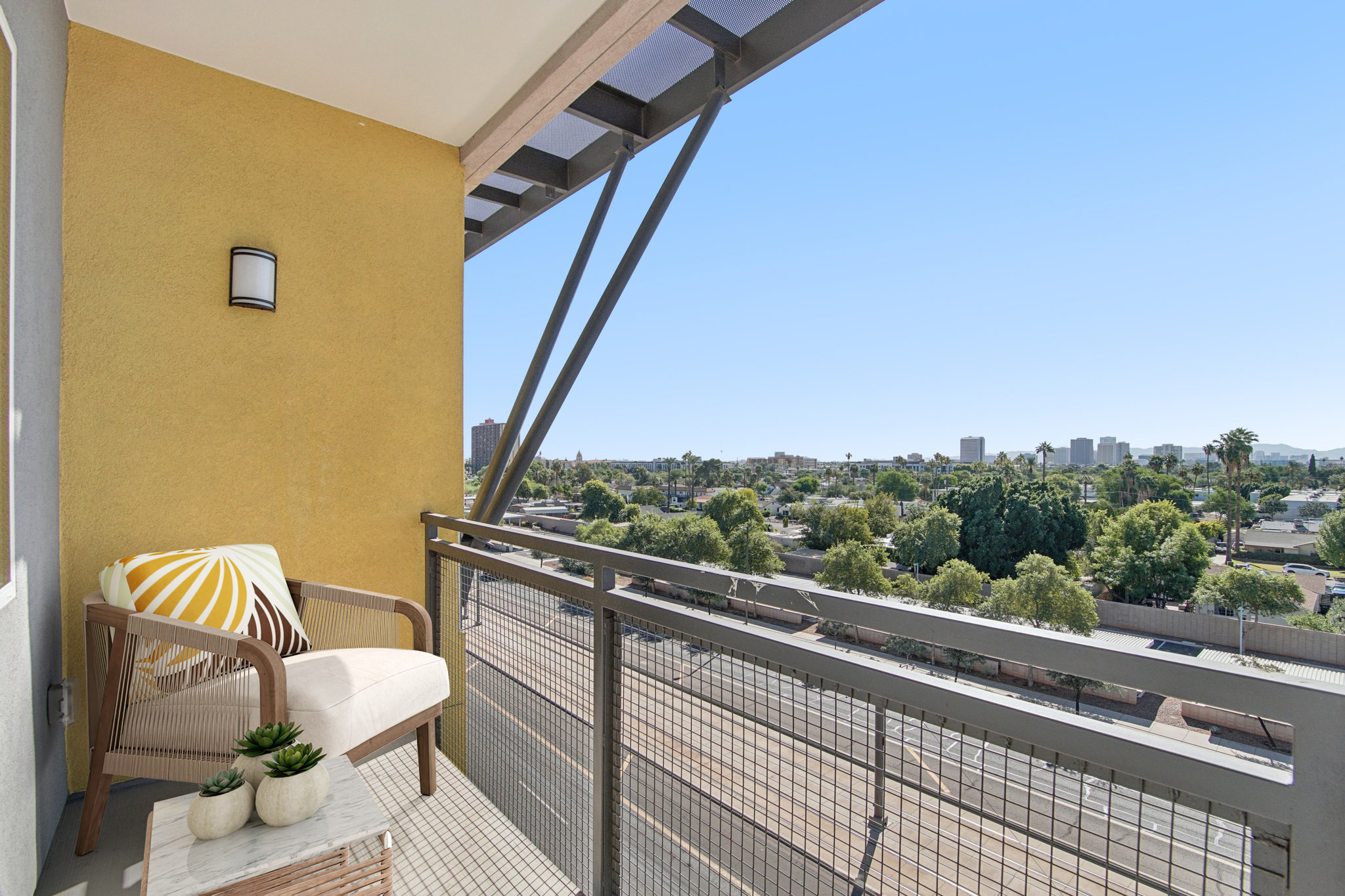 A balcony with a modern chair and small table, overlooking a cityscape with trees and buildings in the distance. The wall is painted yellow, and the sky is clear and blue. A potted plant adds a touch of greenery to the setting.