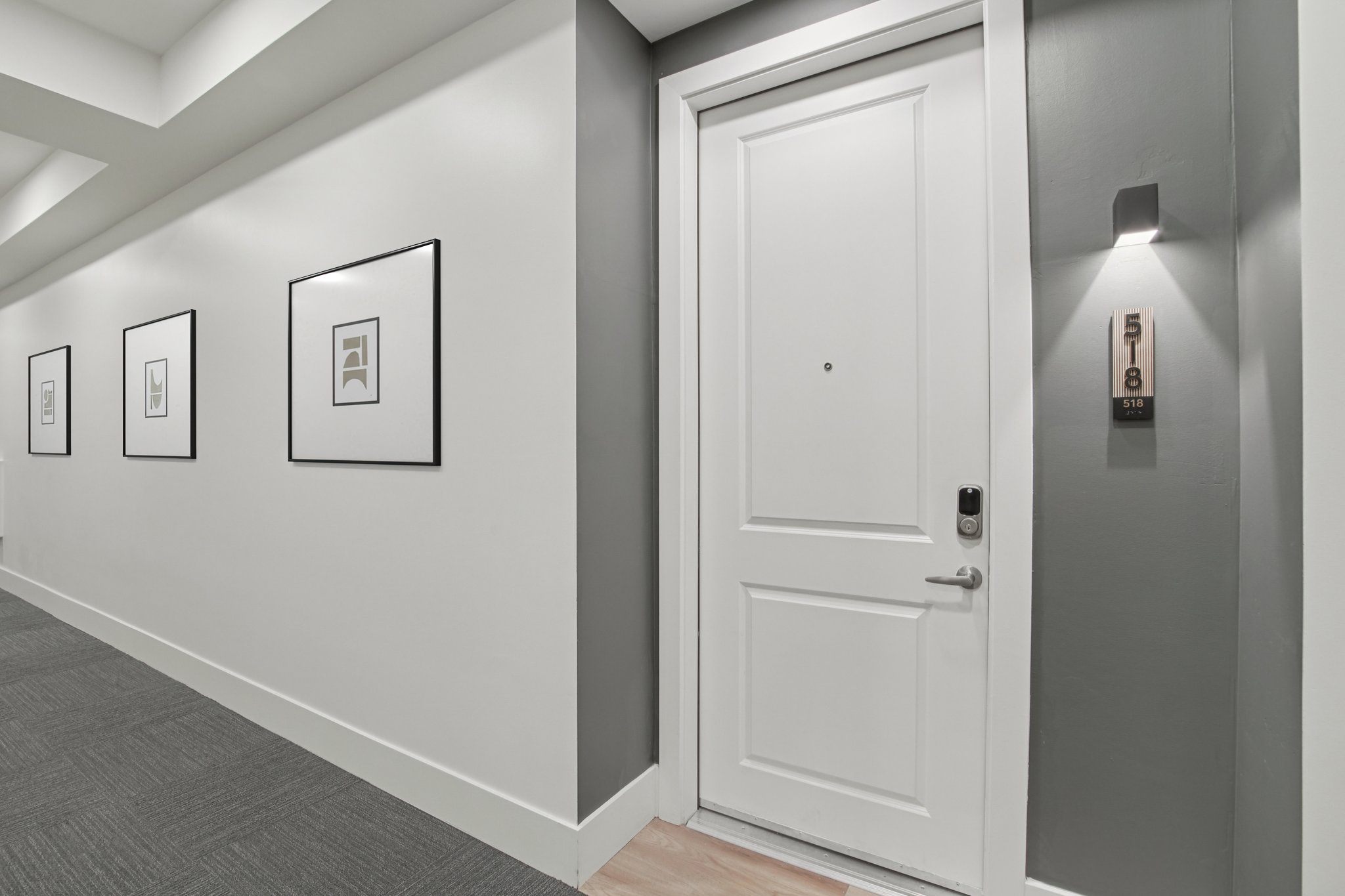 A modern hallway featuring a white door with a handle and a peephole, marked with the number "518." To the left, three framed art pieces adorn the walls, while the wall beside the door is painted gray. Soft lighting highlights the entrance, creating a welcoming atmosphere.