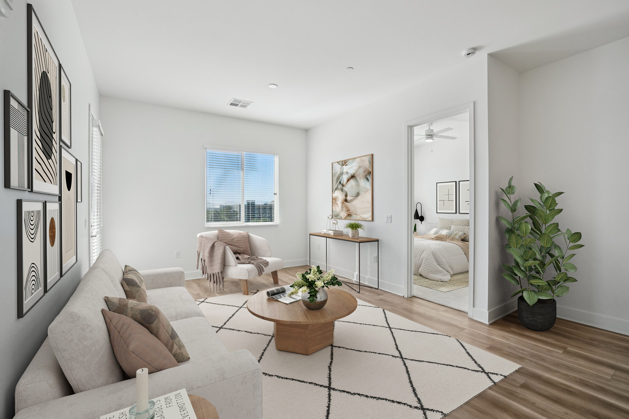 A modern, well-lit living room featuring a light-colored sofa with decorative pillows, a round coffee table, and a textured area rug. A large window lets in natural light, while a potted plant adds a touch of greenery. In the background, there's a doorway leading to a cozy bedroom space.