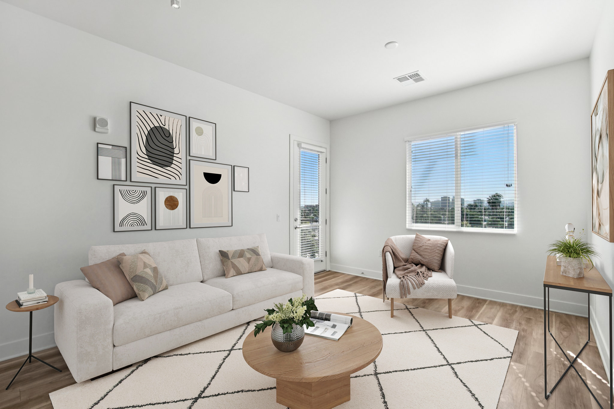 A modern, well-lit living room featuring a light-colored sofa with decorative pillows, a round wooden coffee table, and a small side table. The walls are adorned with abstract art, and a large window provides natural light, while a cozy throw blanket is draped over the sofa. A plant adds a touch of greenery.