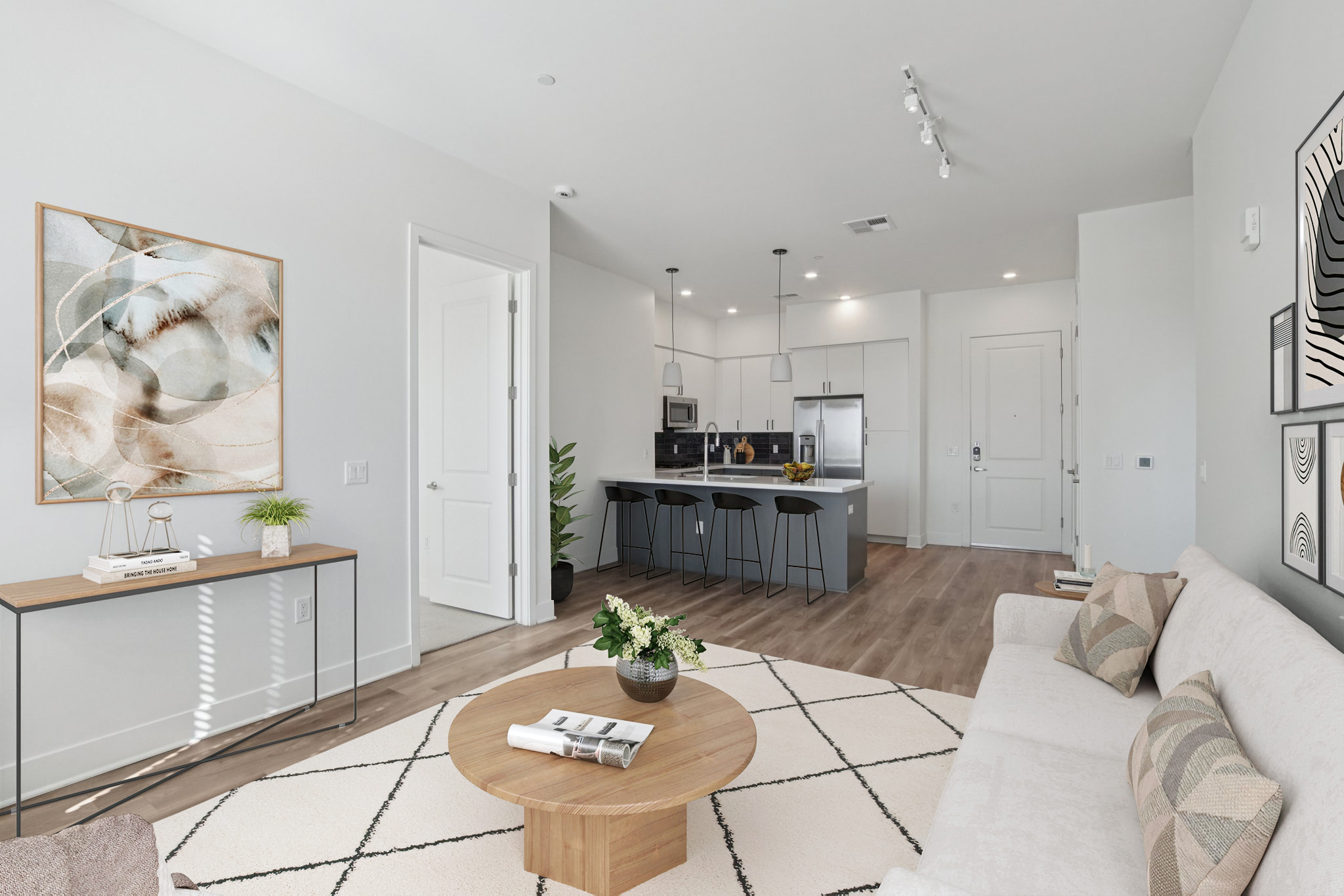 A modern living room featuring a beige sofa with patterned cushions, a round wooden coffee table, and a decorative rug. The space includes a sleek kitchen area with bar stools, contemporary artwork on the walls, and a potted plant, all set in a bright and airy atmosphere.
