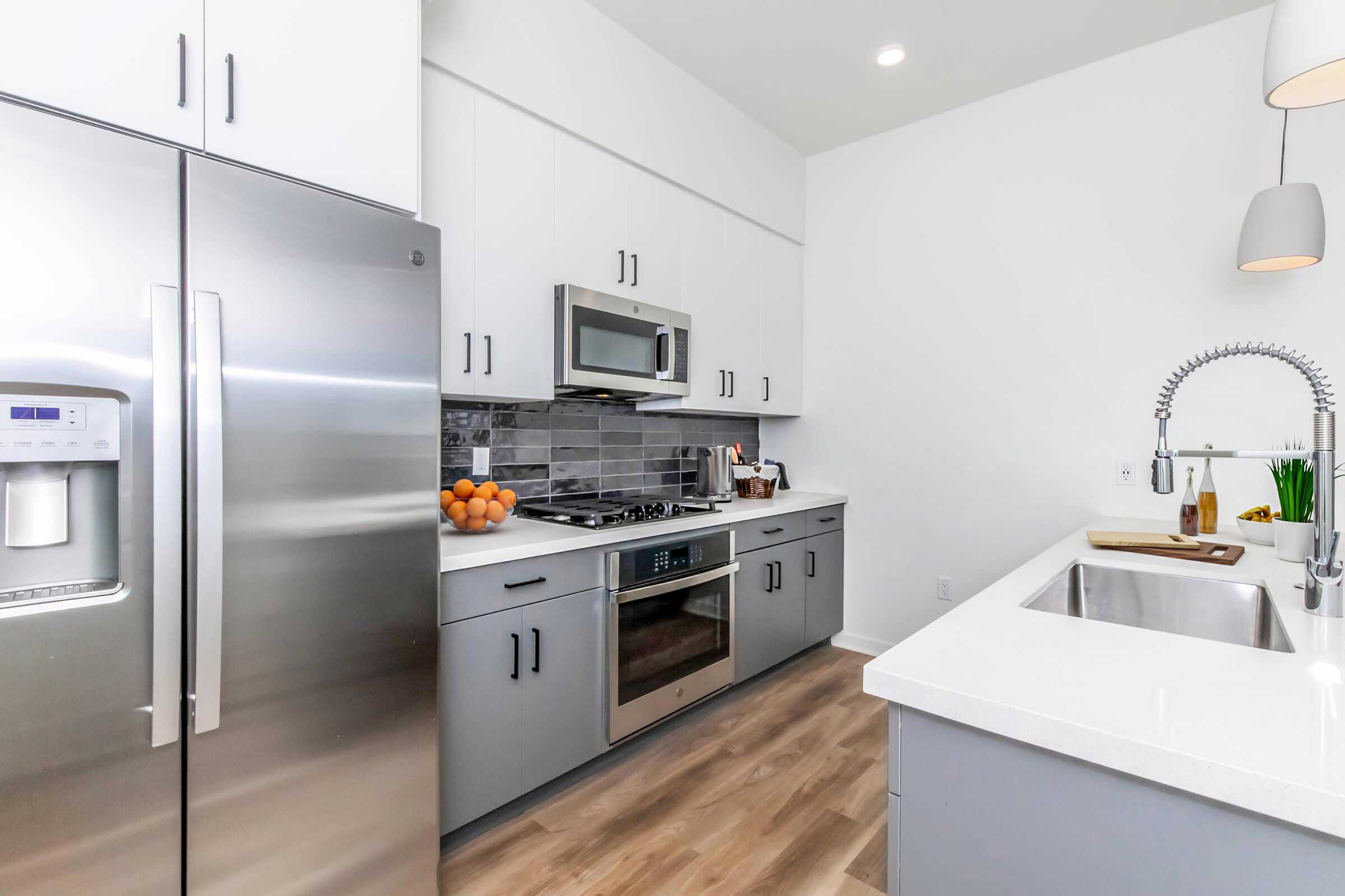 A modern kitchen featuring stainless steel appliances, including a refrigerator, oven, and microwave. The countertops are white with a sleek design, and there is a kitchen island with a sink. A bowl of oranges is on the countertop, and the space is well-lit with natural light.