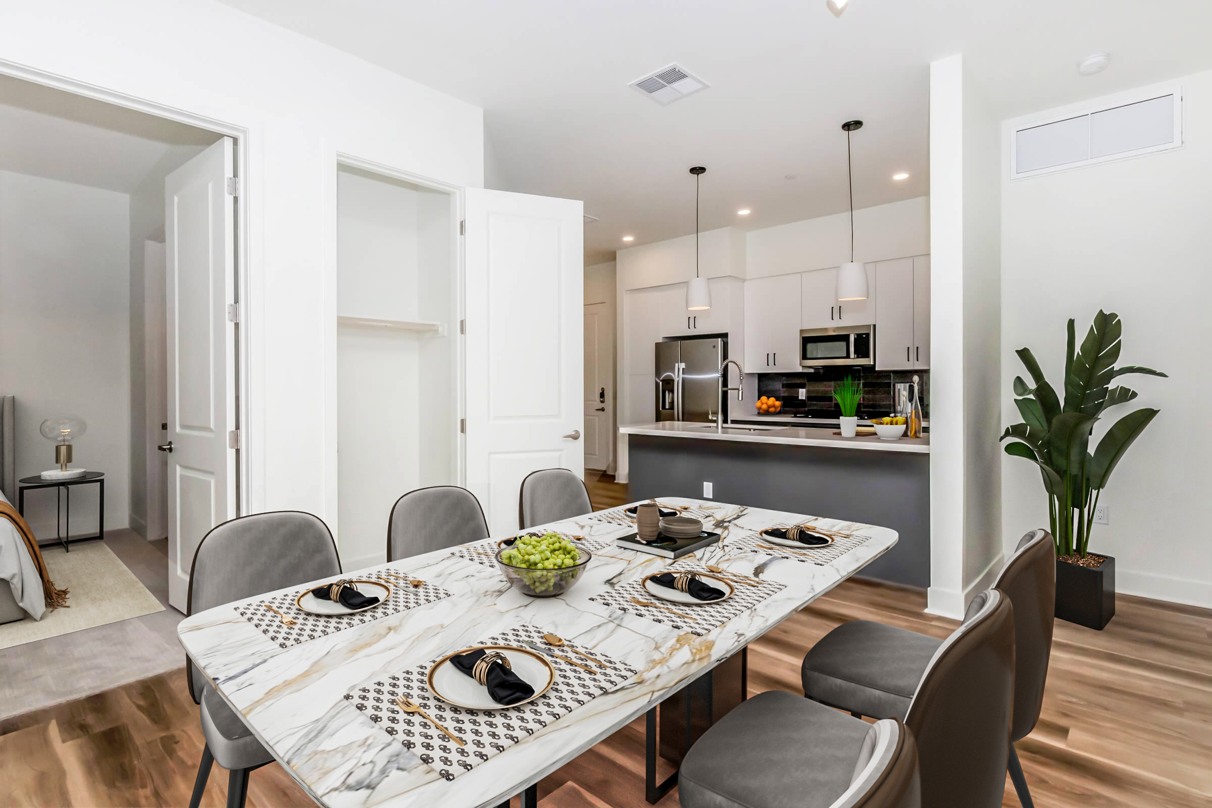 A modern dining area featuring a sleek marble table set with black plates and napkins. The space includes stylish gray chairs and a well-lit kitchen in the background, showcasing stainless steel appliances and a countertop with fresh fruit. A large green plant adds a touch of nature to the room.