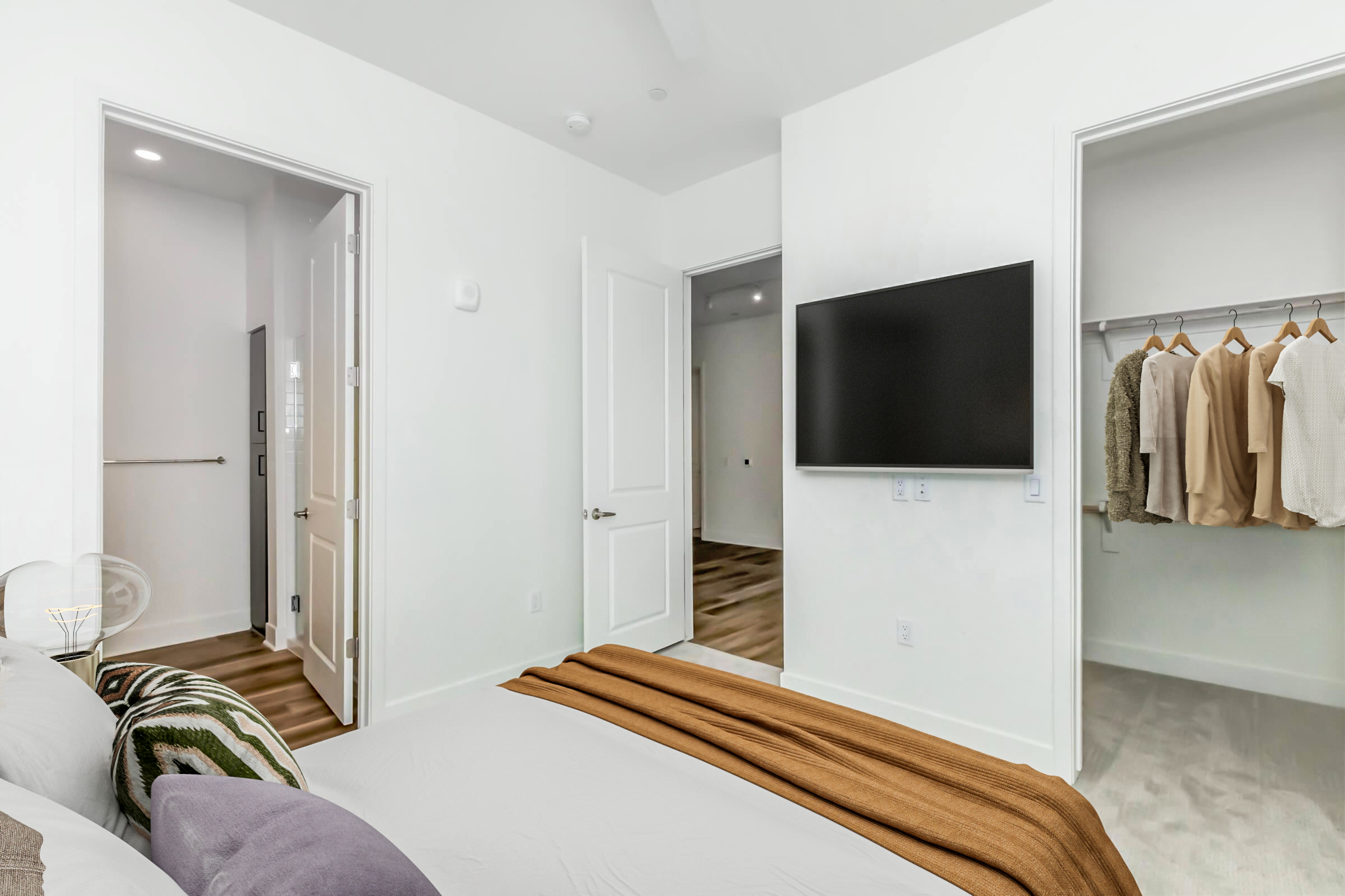 A modern bedroom featuring a bed with a neutral-colored blanket, a wall-mounted TV, and a closet with hanging clothes. There are two doors leading to adjacent rooms. The walls are painted white, and the flooring is light wood. The decor is simple and contemporary.
