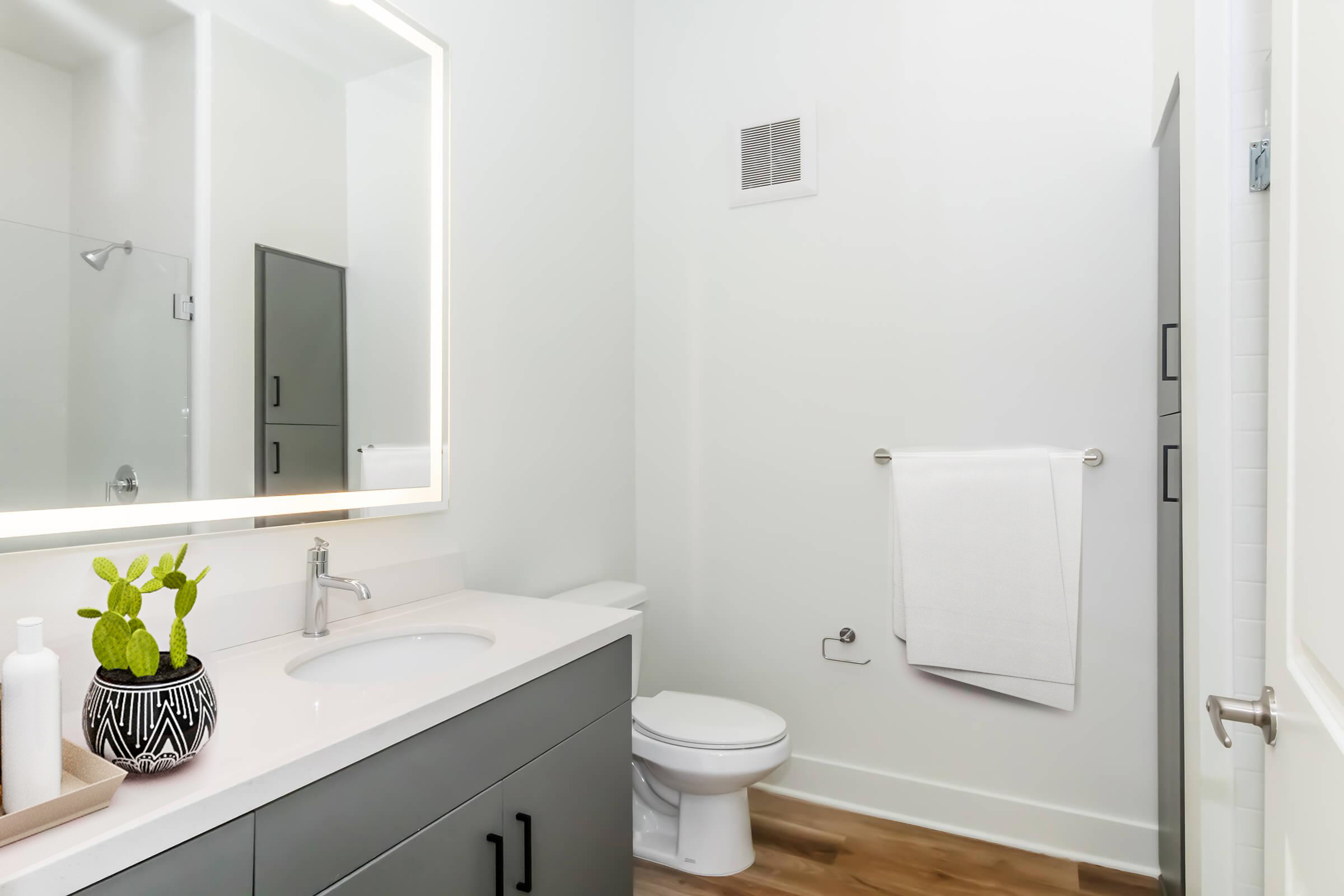 A modern bathroom featuring a sleek gray vanity with a white sink, a round mirror illuminated by LED lighting, a toilet, and a walk-in shower. A small potted plant and a white towel hang on a rack, with wooden flooring enhancing the contemporary design.