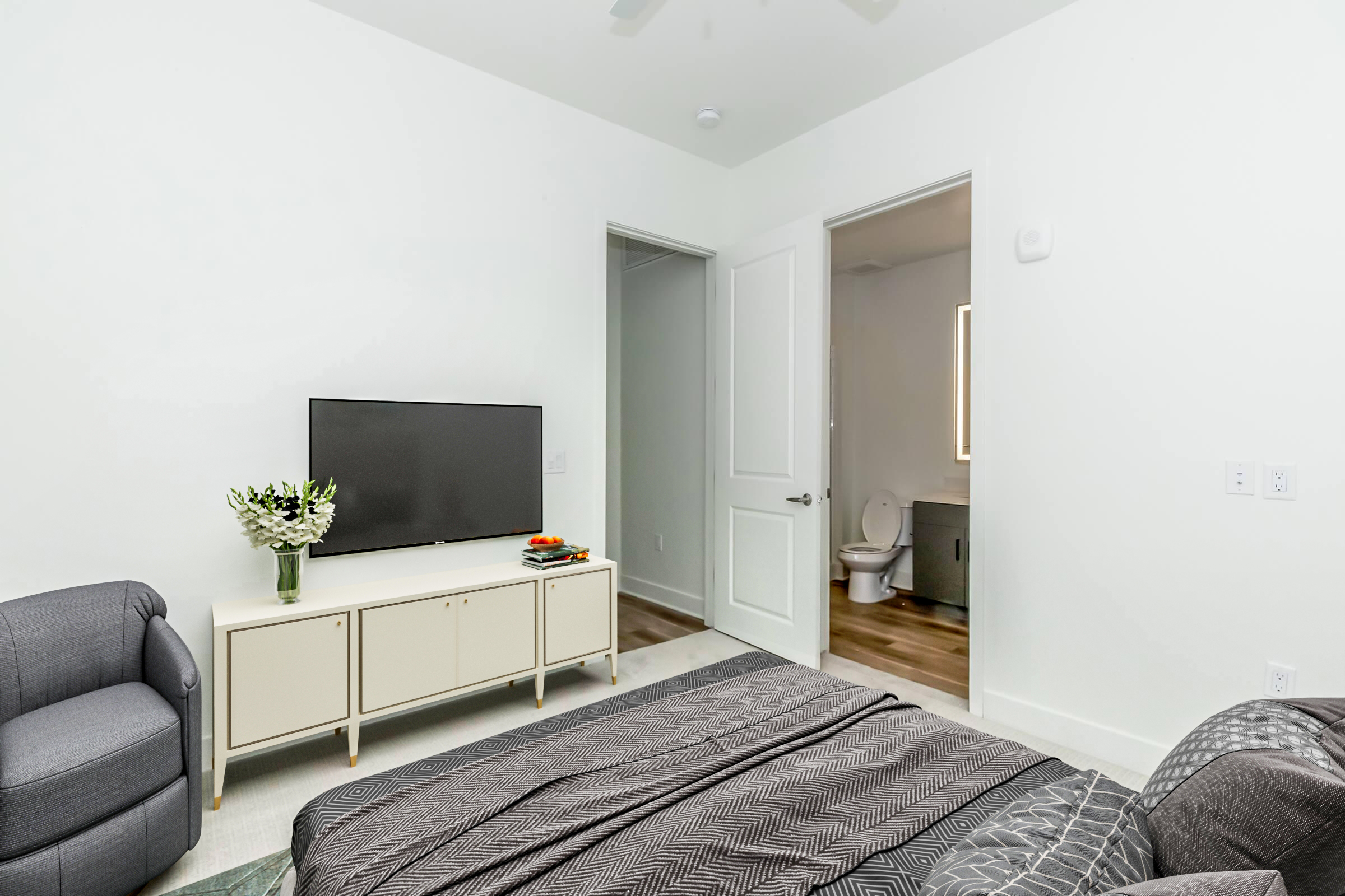 A modern bedroom featuring a plush gray bedspread and an armchair. A TV is mounted on a white media console with a vase of flowers on top. A doorway leads to a bathroom with a visible toilet and a mirror. The walls are painted white, and the flooring includes light wood accents.
