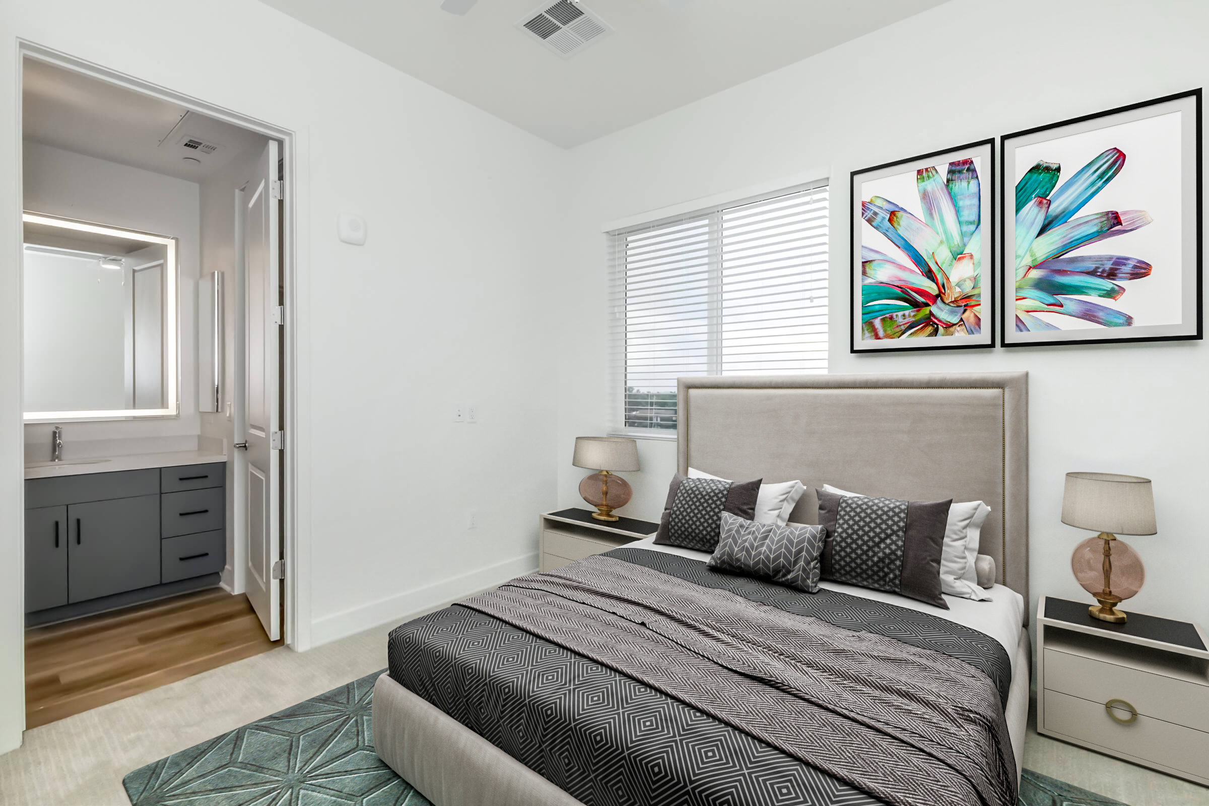 A modern bedroom featuring a queen-sized bed with gray bedding and decorative pillows. The room includes a nightstand with lamps on either side, colorful artwork on the wall, and an adjoining bathroom visible through an open door. The space has white walls and a textured area rug.