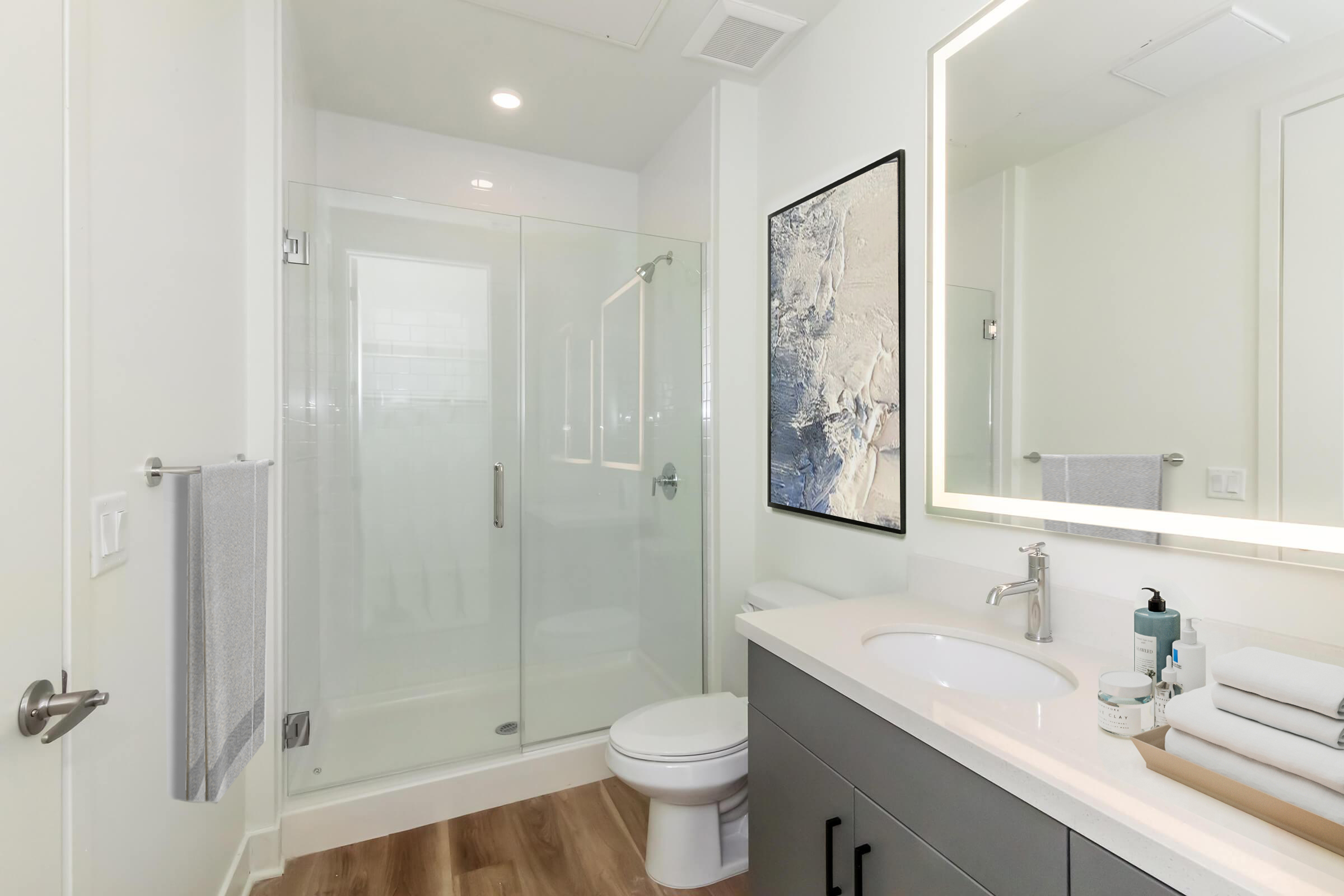 Modern bathroom featuring a glass shower enclosure, a sleek vanity with a light-colored sink, a large round mirror illuminated by ambient lighting, and a large piece of artwork on the wall. There are towels hanging, toiletries on the counter, and warm wooden flooring.