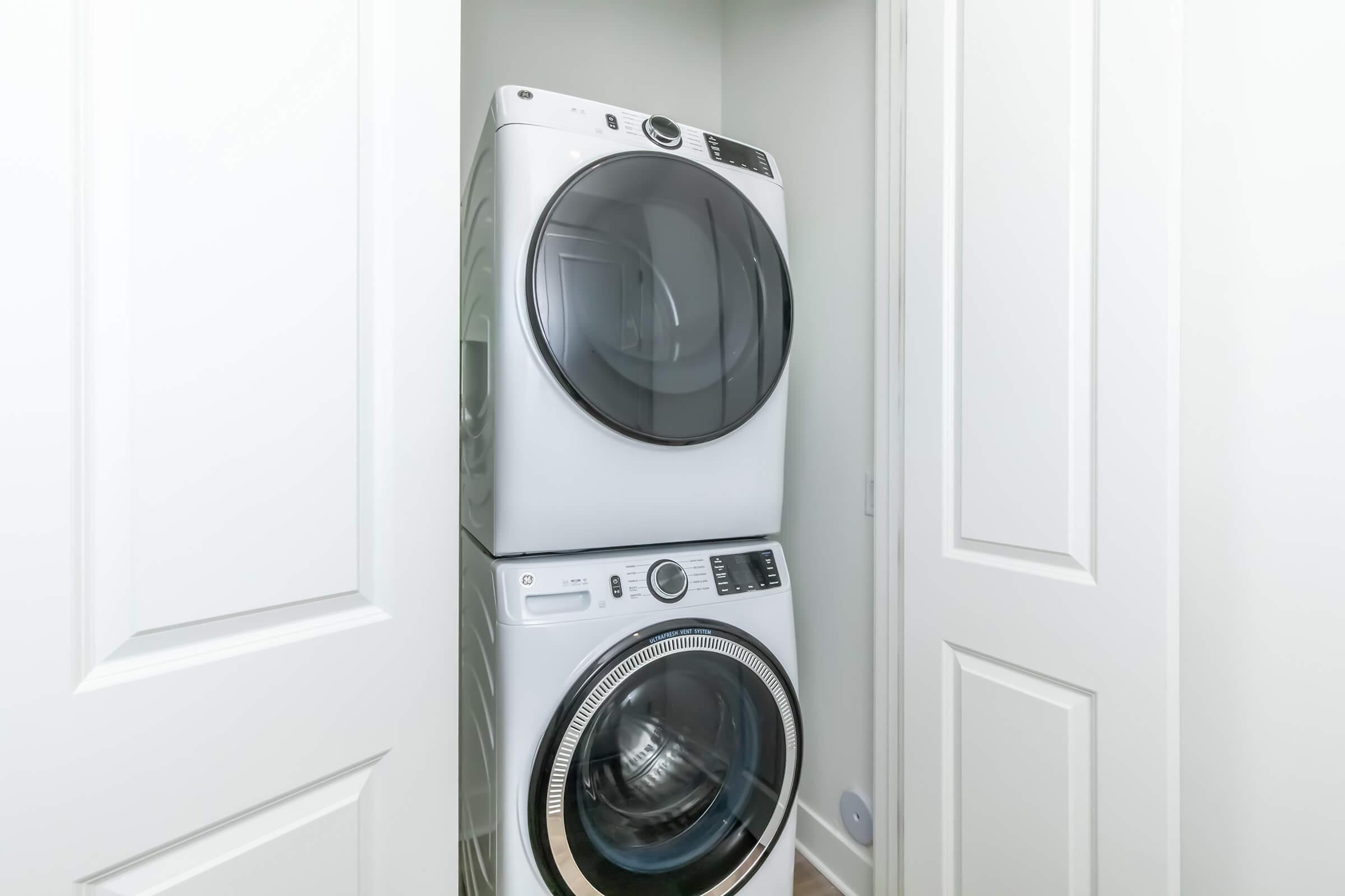 A stackable washer and dryer located in a small closet. The appliances are modern with a sleek design, featuring large glass doors and control panels on the front. The closet has white doors, providing a neat and organized appearance.