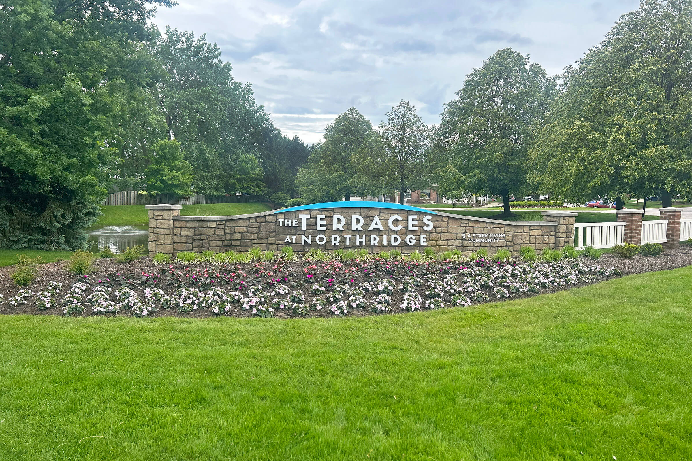 A stone sign reading "The Terraces at Northridge" surrounded by lush greenery and colorful flower beds. The scene features a cloudy sky and well-maintained landscaping, creating an inviting atmosphere.