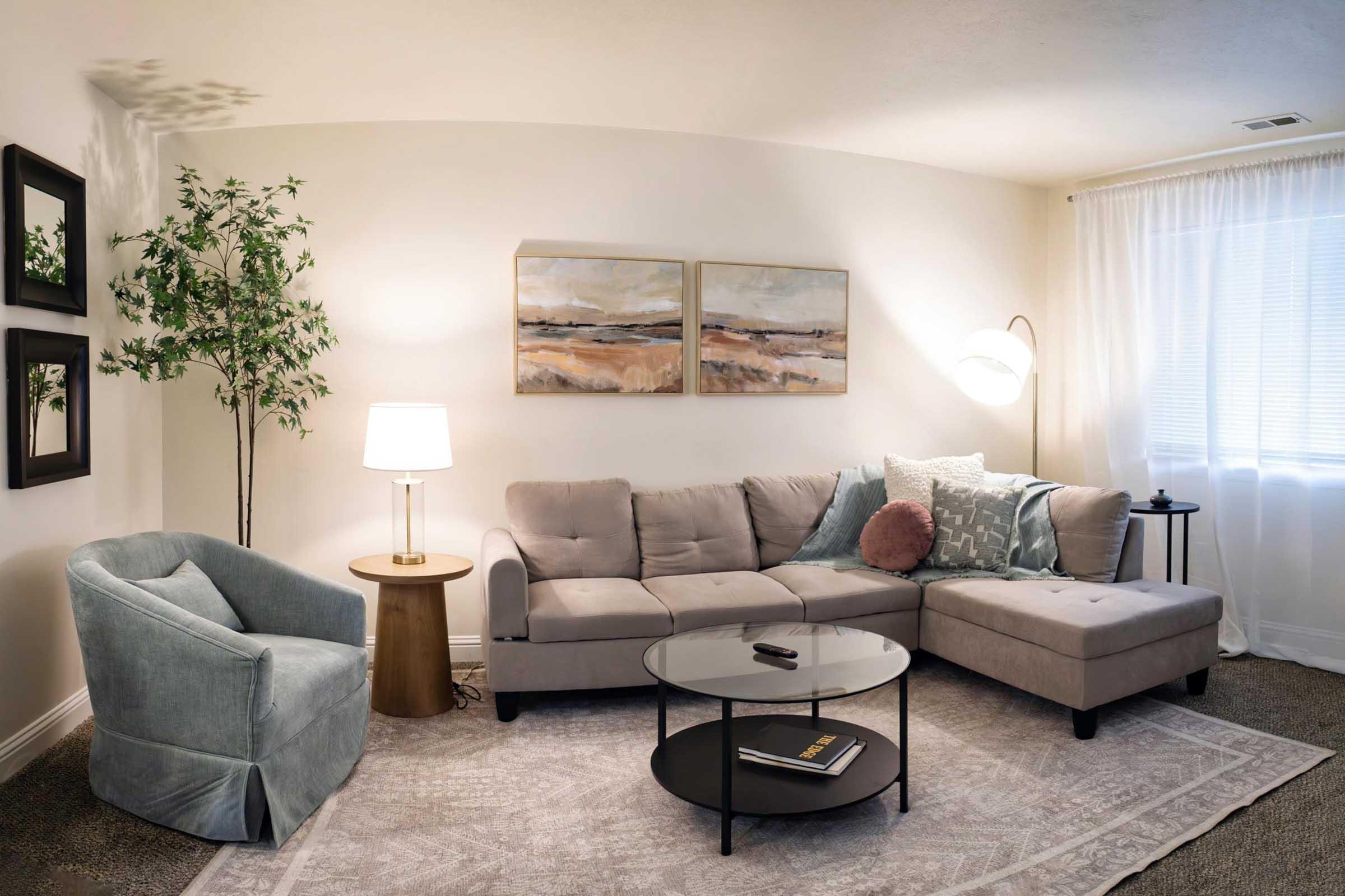 A cozy living room featuring a light gray sectional sofa with decorative pillows, a round glass coffee table, and a stylish armchair. A floor lamp illuminates the space beside a small side table. The walls are adorned with two abstract paintings, and a greenery plant adds a touch of nature to the decor.