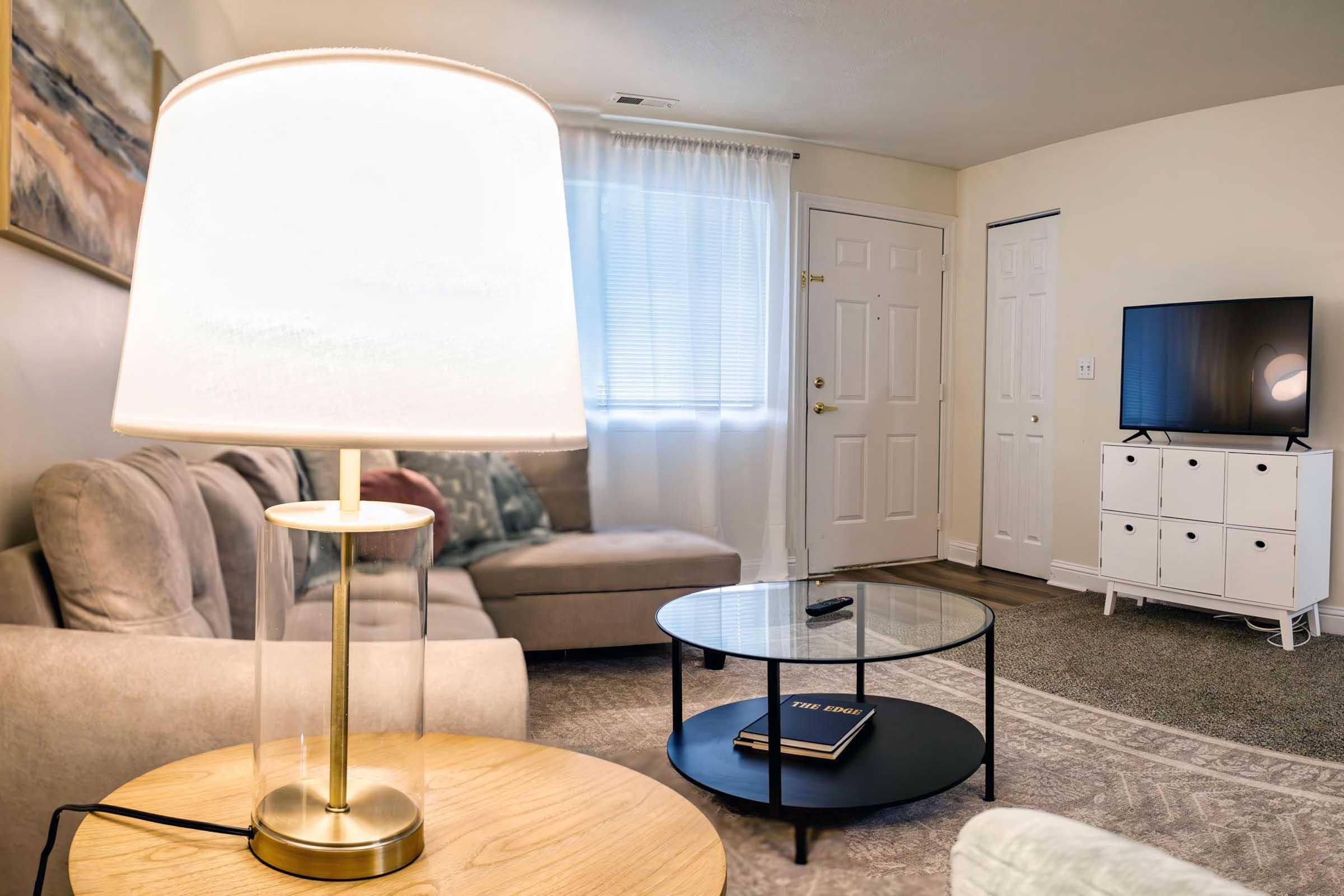A cozy living room featuring a beige sofa, a round black coffee table with books, a lamp on a wooden side table, and a white media console with a TV. Light filters through a sheer curtain, adding warmth to the space. The decor is simple and inviting, creating a comfortable atmosphere.