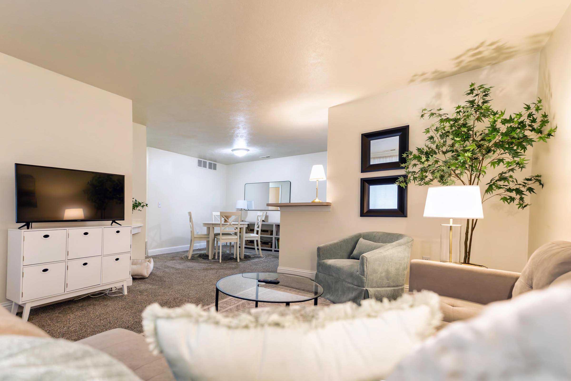A cozy living room featuring a light-colored sofa with decorative pillows, a glass coffee table, and a flat-screen TV on a white modern cabinet. There is a dining area in the background with a white table and chairs, and a floor lamp. A tall plant adds a touch of greenery to the space. The walls are painted in neutral tones.